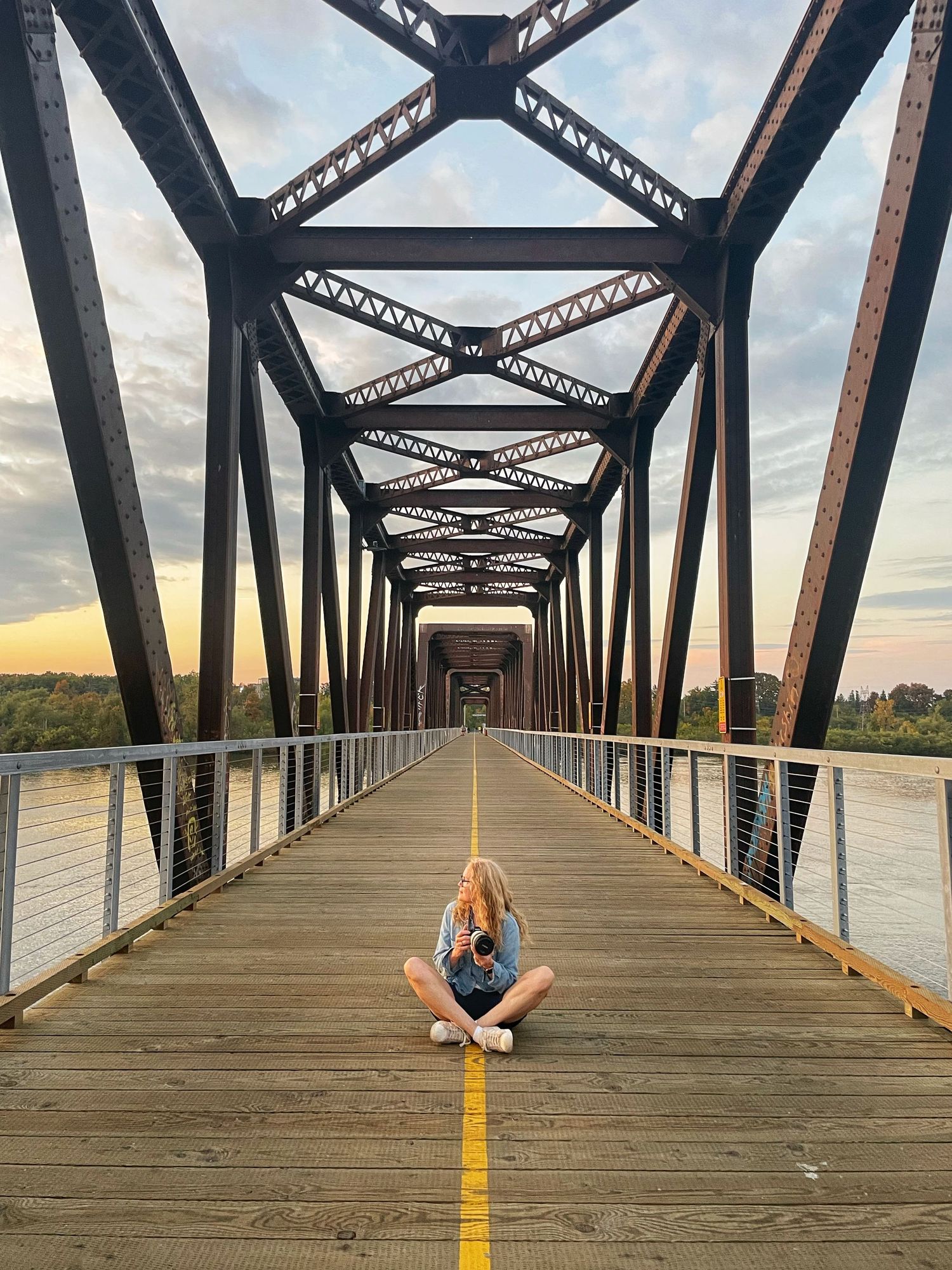 Long wooden bridge at sunset with a photographer sitting crosslegged ,looking away towards the sunset.