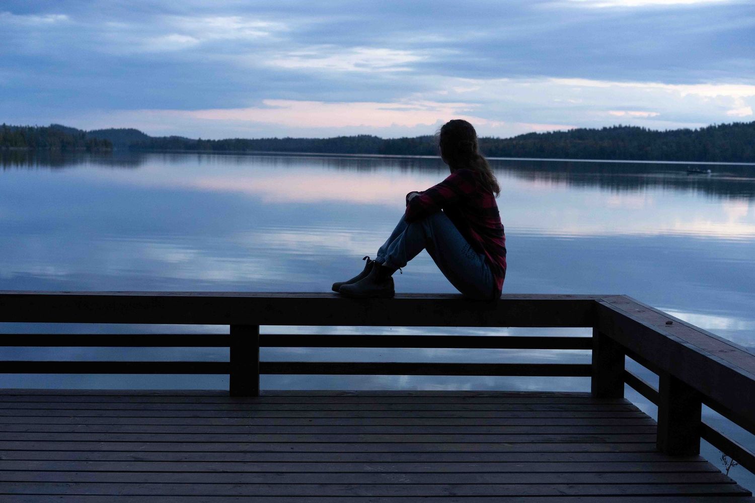 Silhouetted figure perches on a wooden railing viewing calm lake waters at dusk.