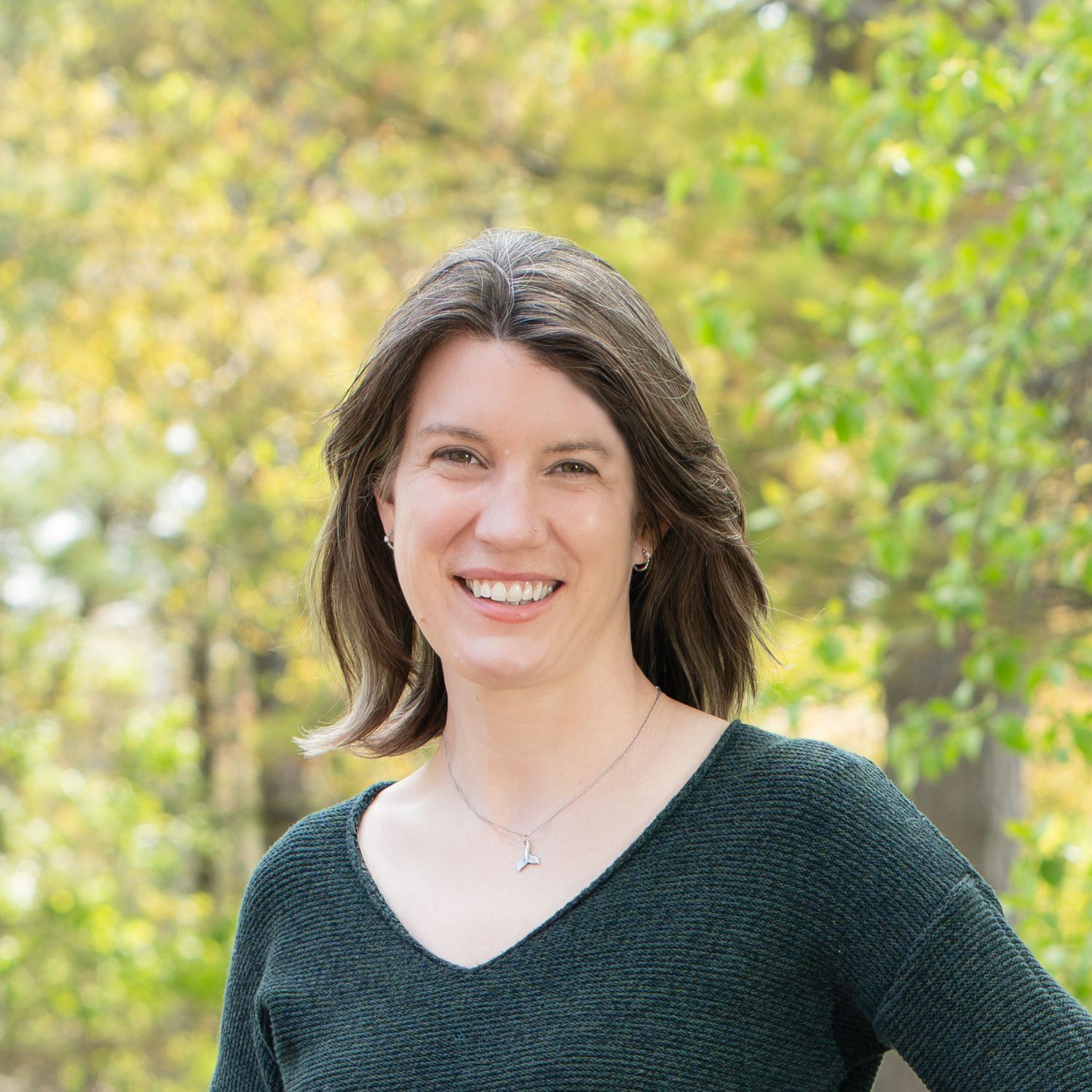 Smiling woman with brown hair wearing a dark green sweater outdoors with blurred green trees in background.
