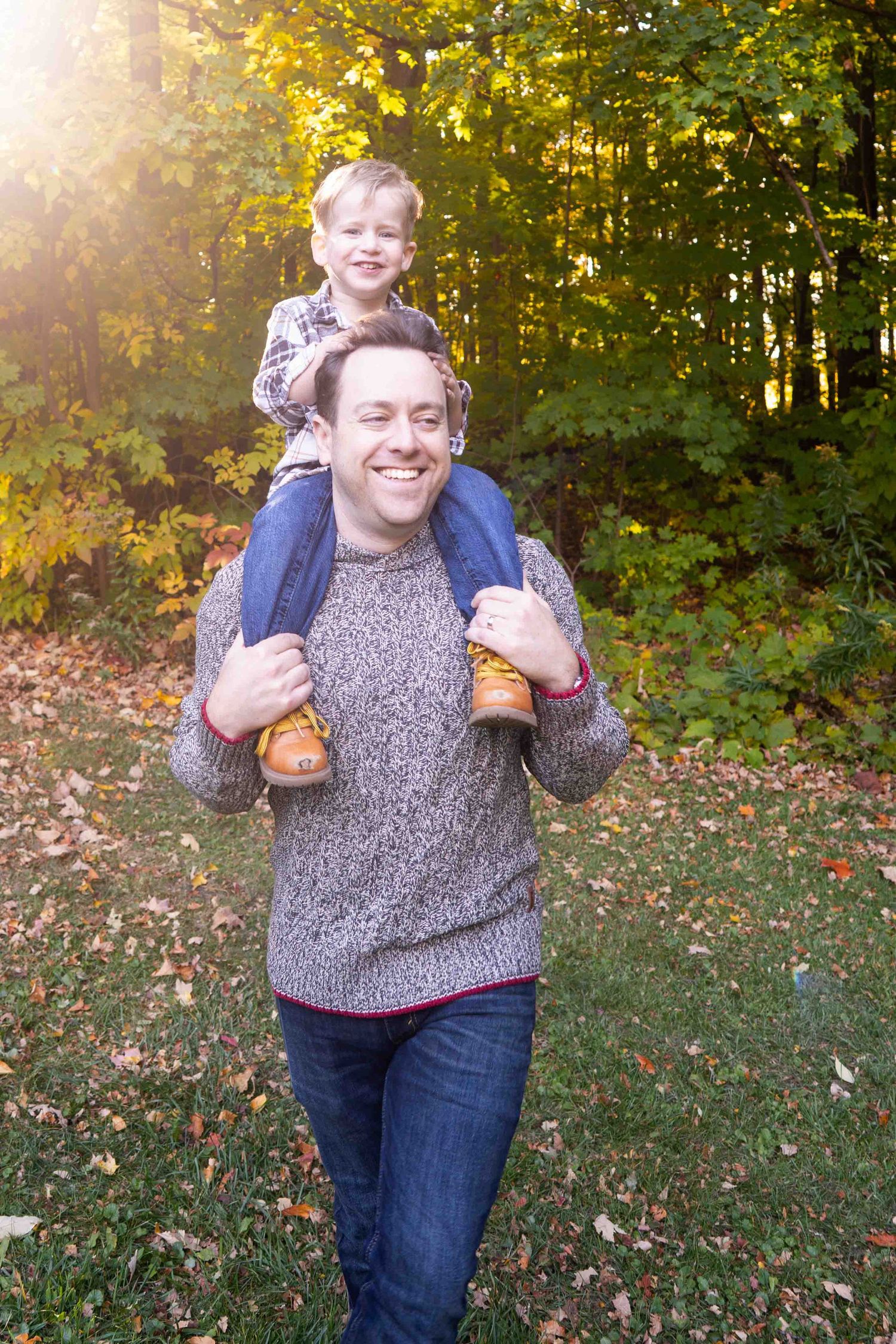 Someone wearing a gray sweater poses playfully outdoors with his son on his shoulder during fall with golden leaves in background.