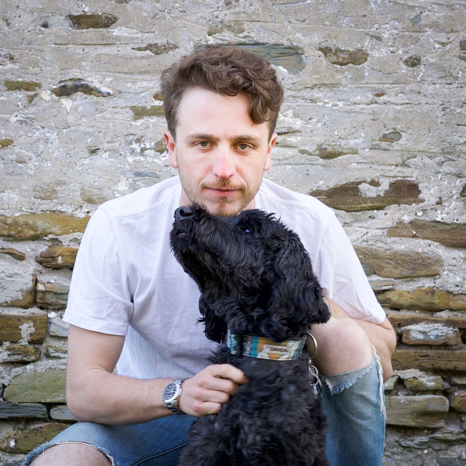 A  black dog sits with its owner against a stone wall background.