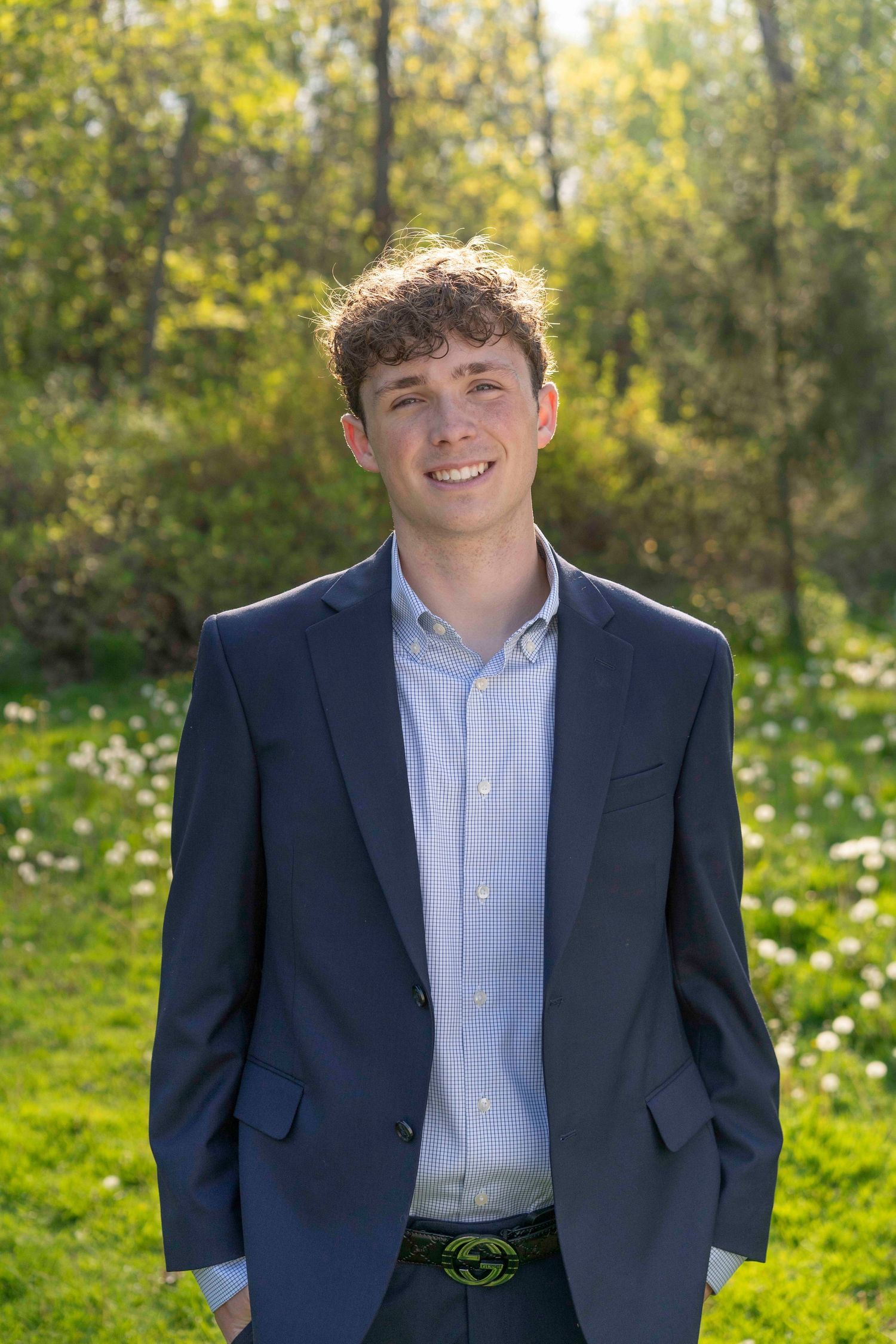 A person in a navy suit and light blue shirt poses outdoors against a backdrop of autumn trees and greenery.