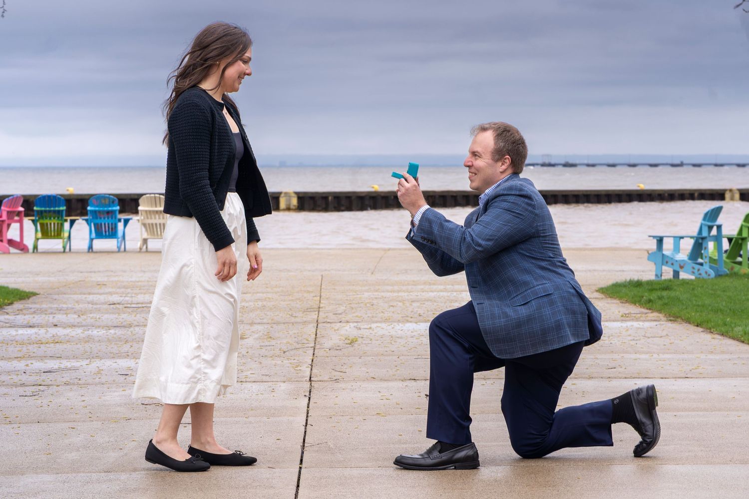 Man on one knee proposing with ring box to woman by lakeside with colorful Adirondack chairs in background.