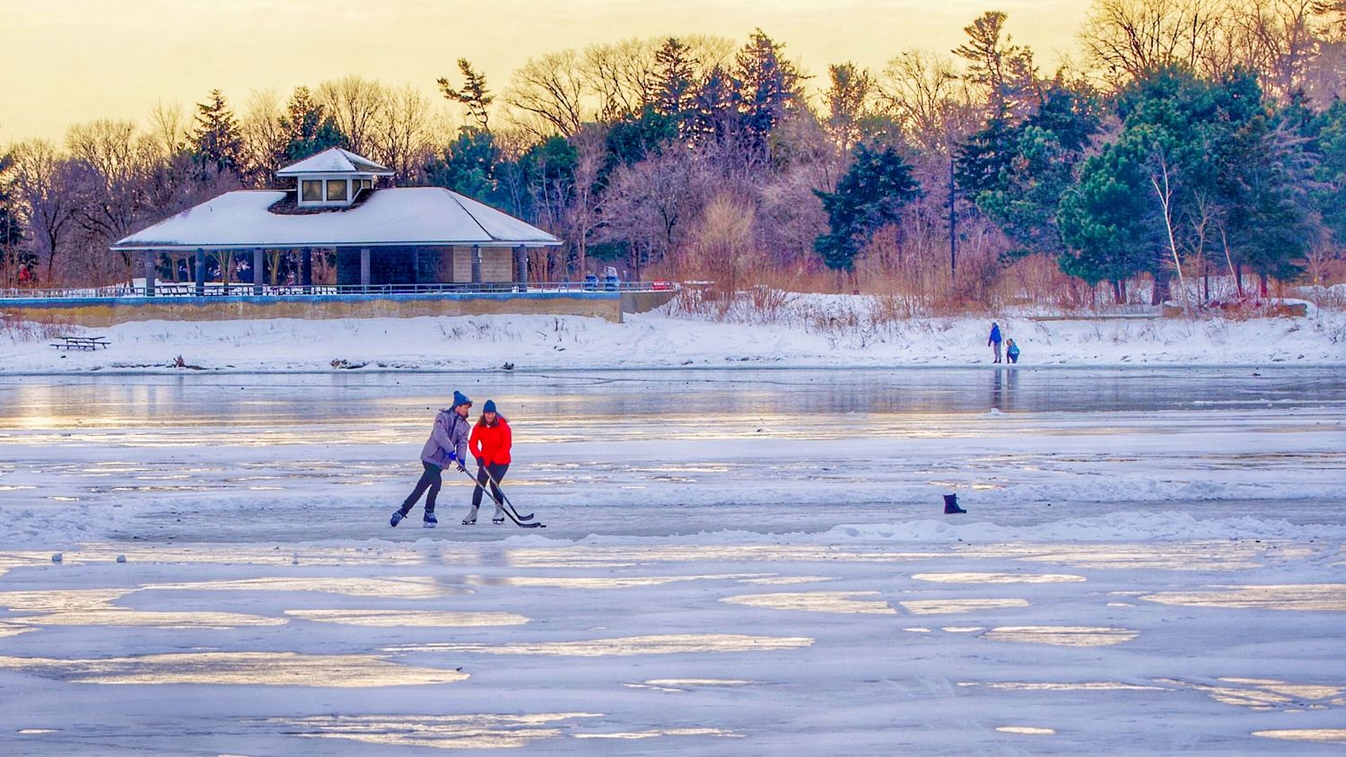 People ice skating on a frozen lake at sunset with trees in the background.