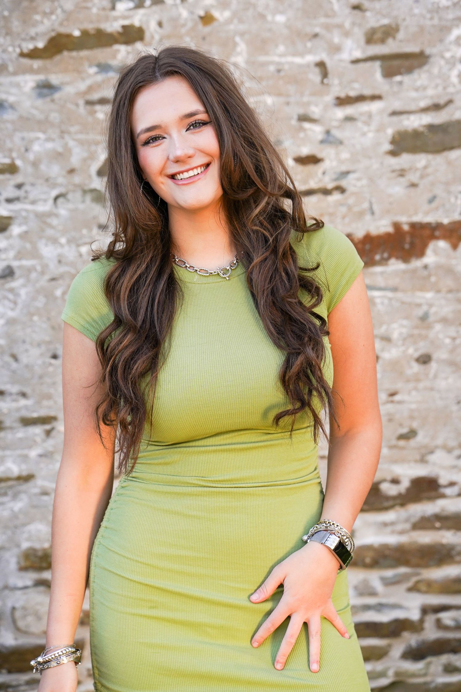 Young woman with long brown hair smiling in a green dress, wearing a silver necklace and bracelet, posed against a stone wall.