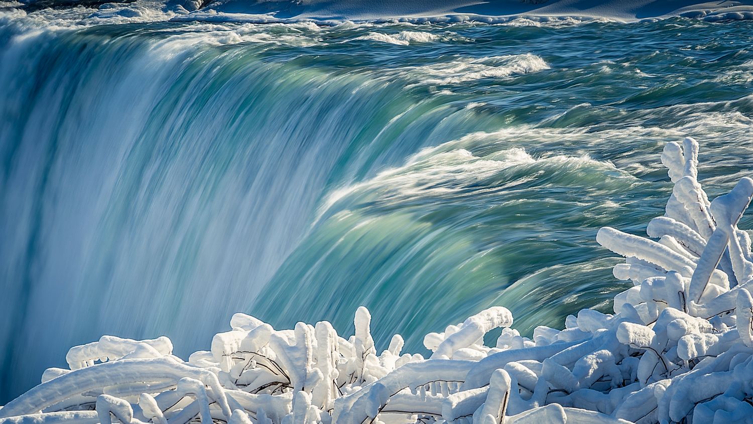 Frozen Niagara Falls in winter with turquoise rushing water cascading over icy snow-covered rocks in stunning detail.