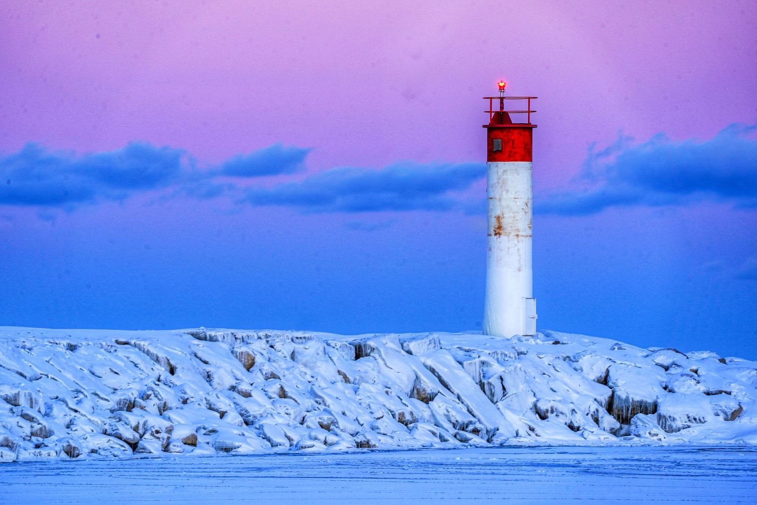 A lighthouse stands at the end of a snow-covered breakwater against a vibrant purple and blue winter sky.