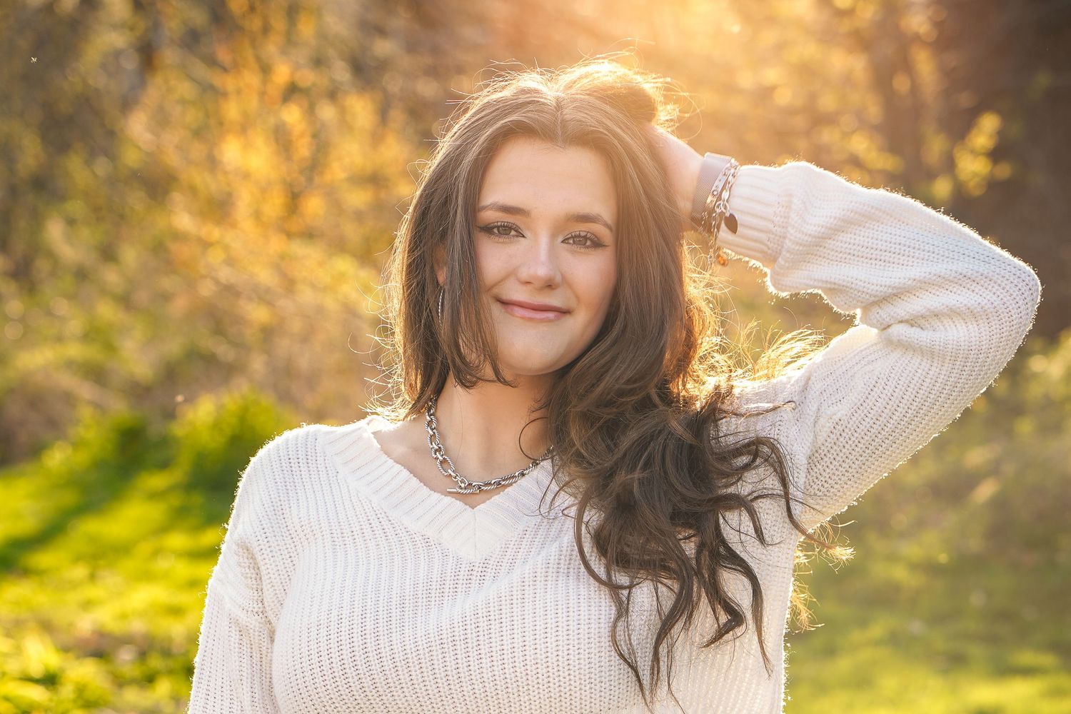 A person in a white knit sweater poses outdoors during golden hour with sunlit autumn foliage in the background.