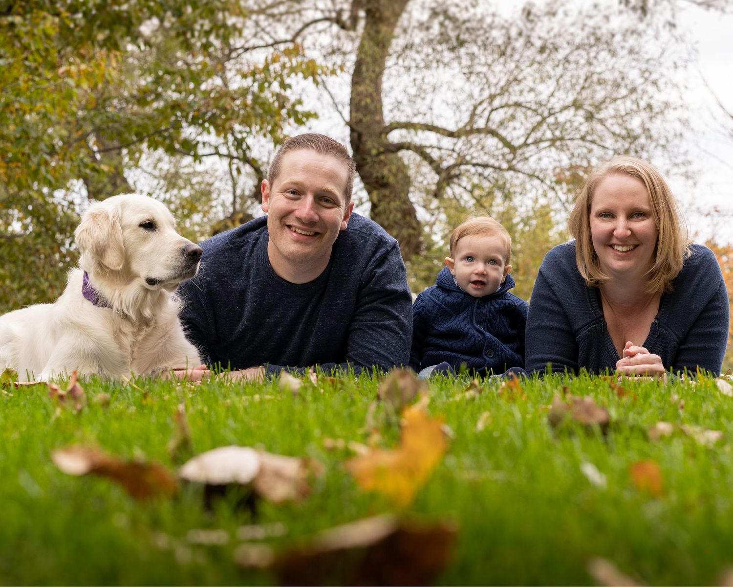 A family and their golden retriever dog lying in grass with autumn leaves during a fall outdoor photoshoot.