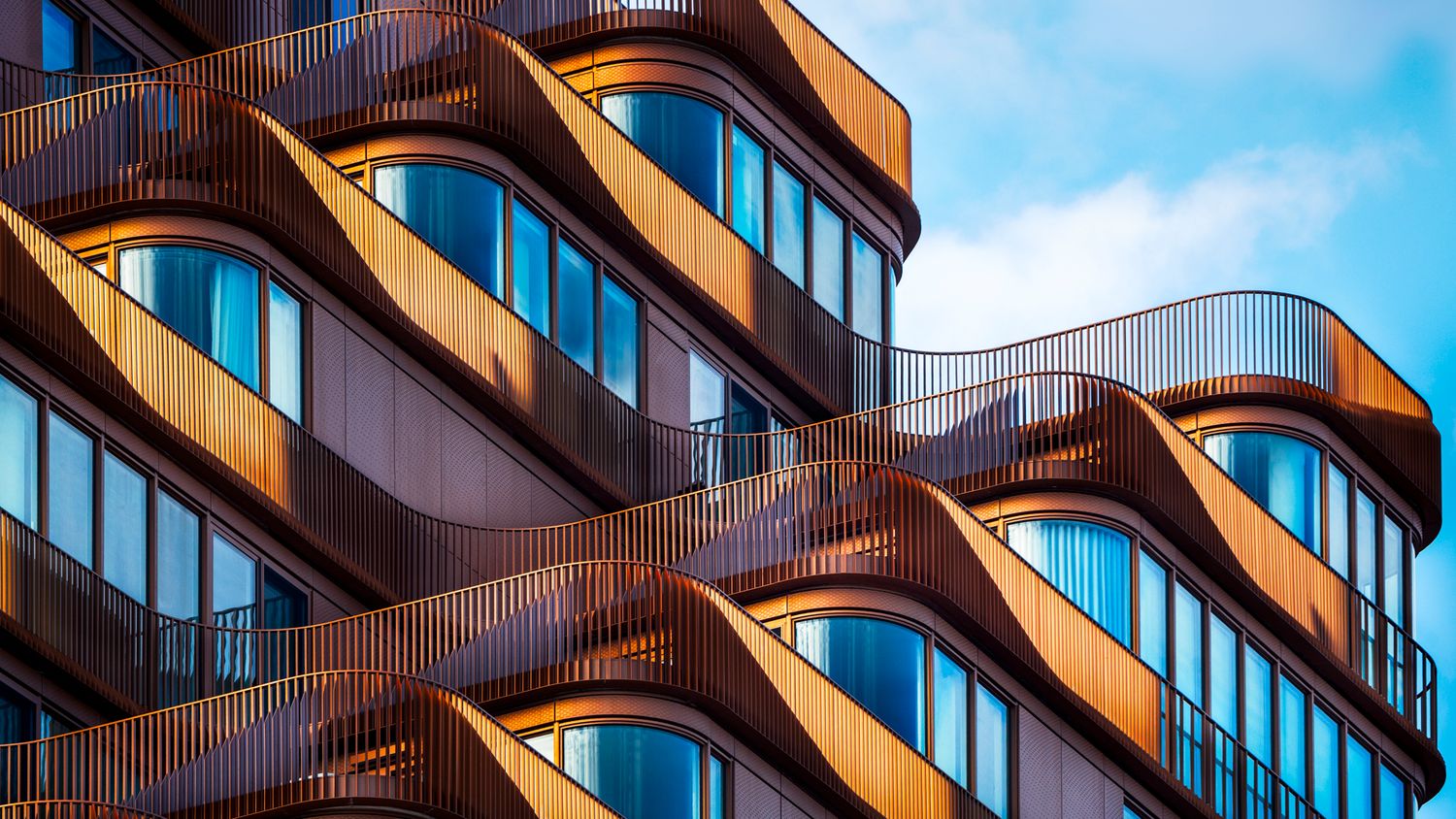 Modern building facade with wavy copper balconies and curved blue glass windows against a partly cloudy sky.