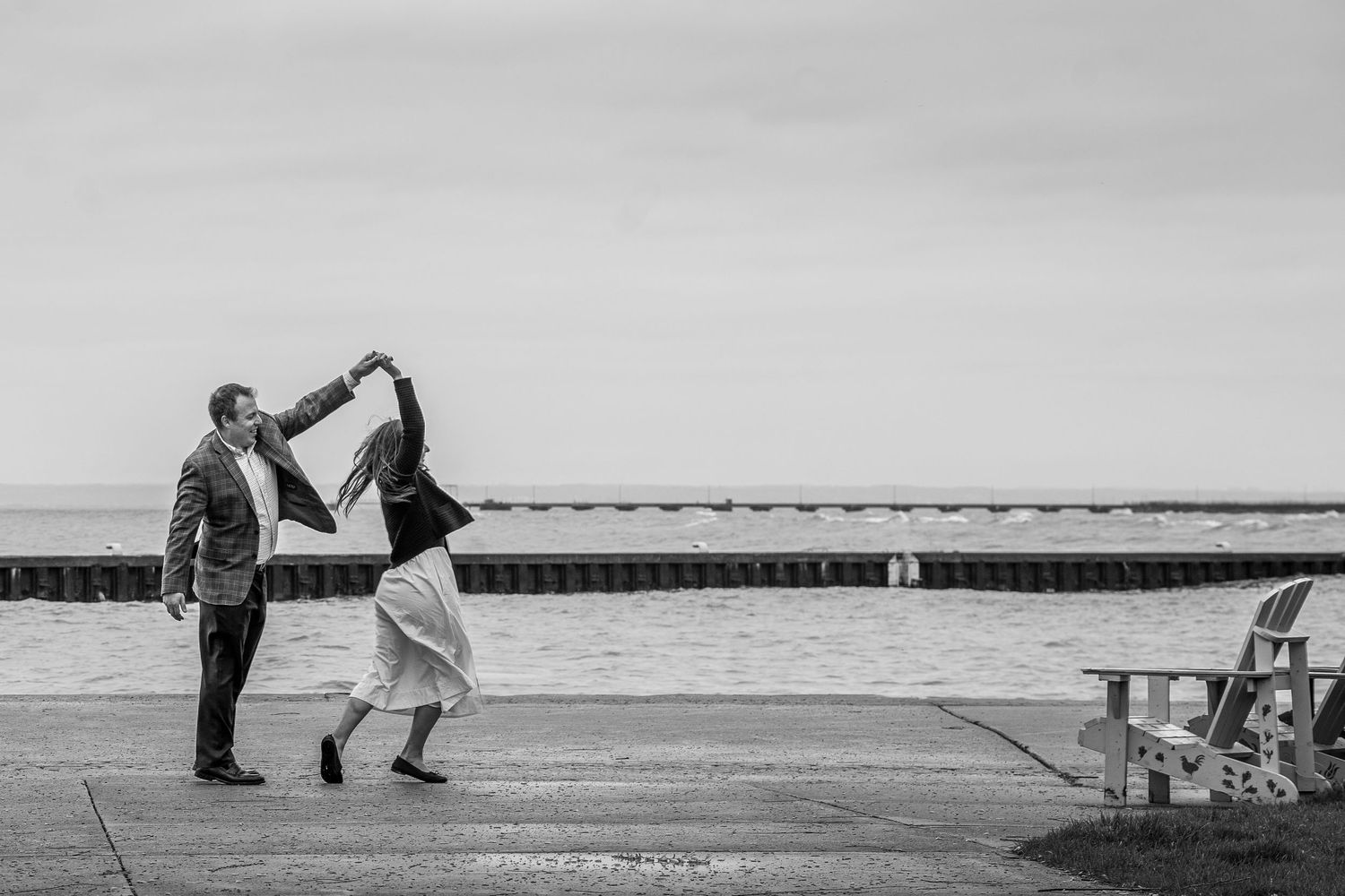 Black and white photo of a couple dancing joyfully by the waterfront, man spinning woman in elegant attire near a bench.