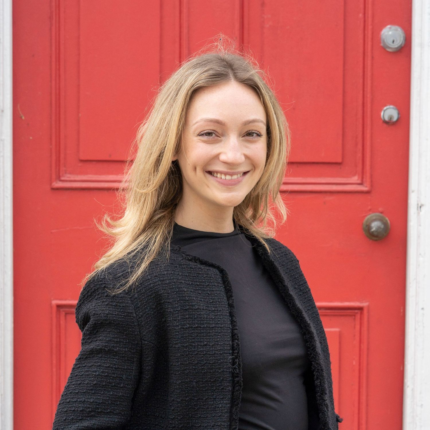 Professional wearing a black coat stands smiling against a bright red door background.