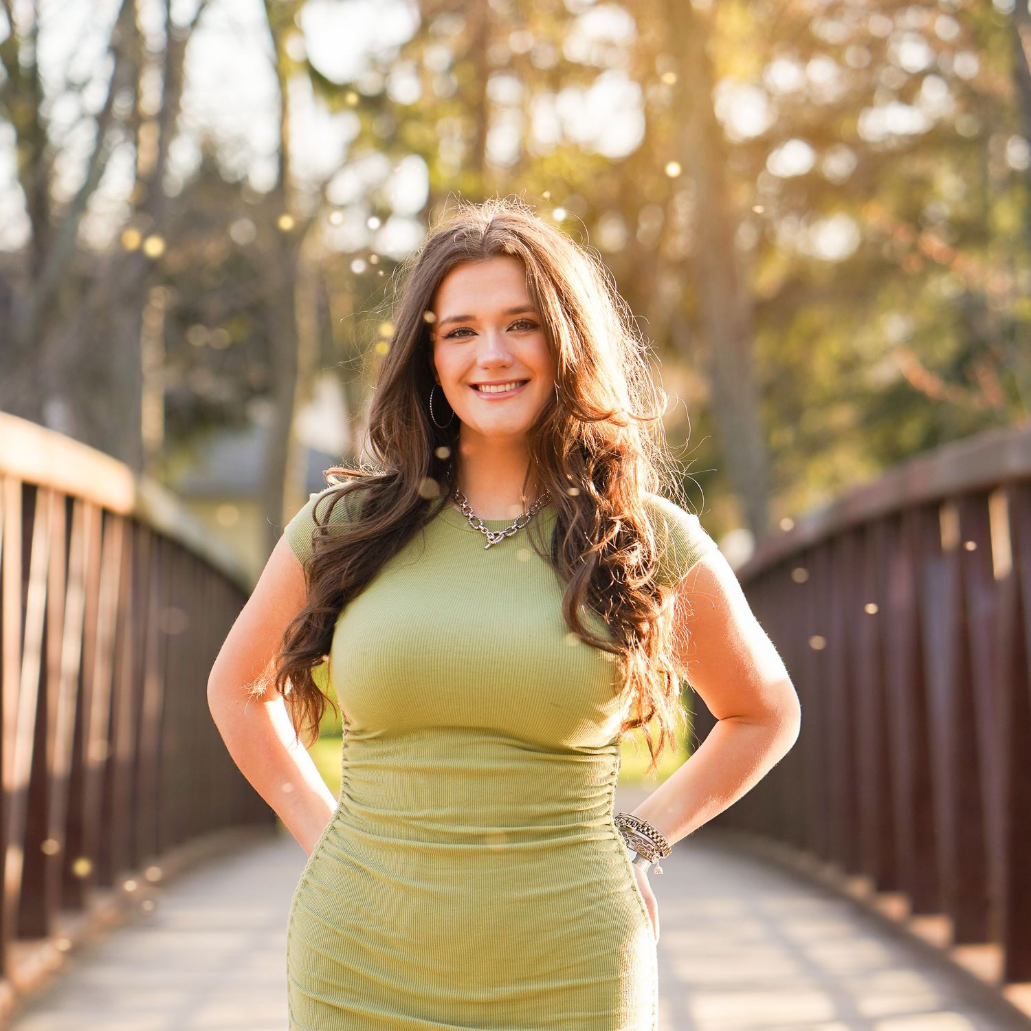 Person in a lime green top poses outdoors on a wooden bridge during golden hour sunset lighting.