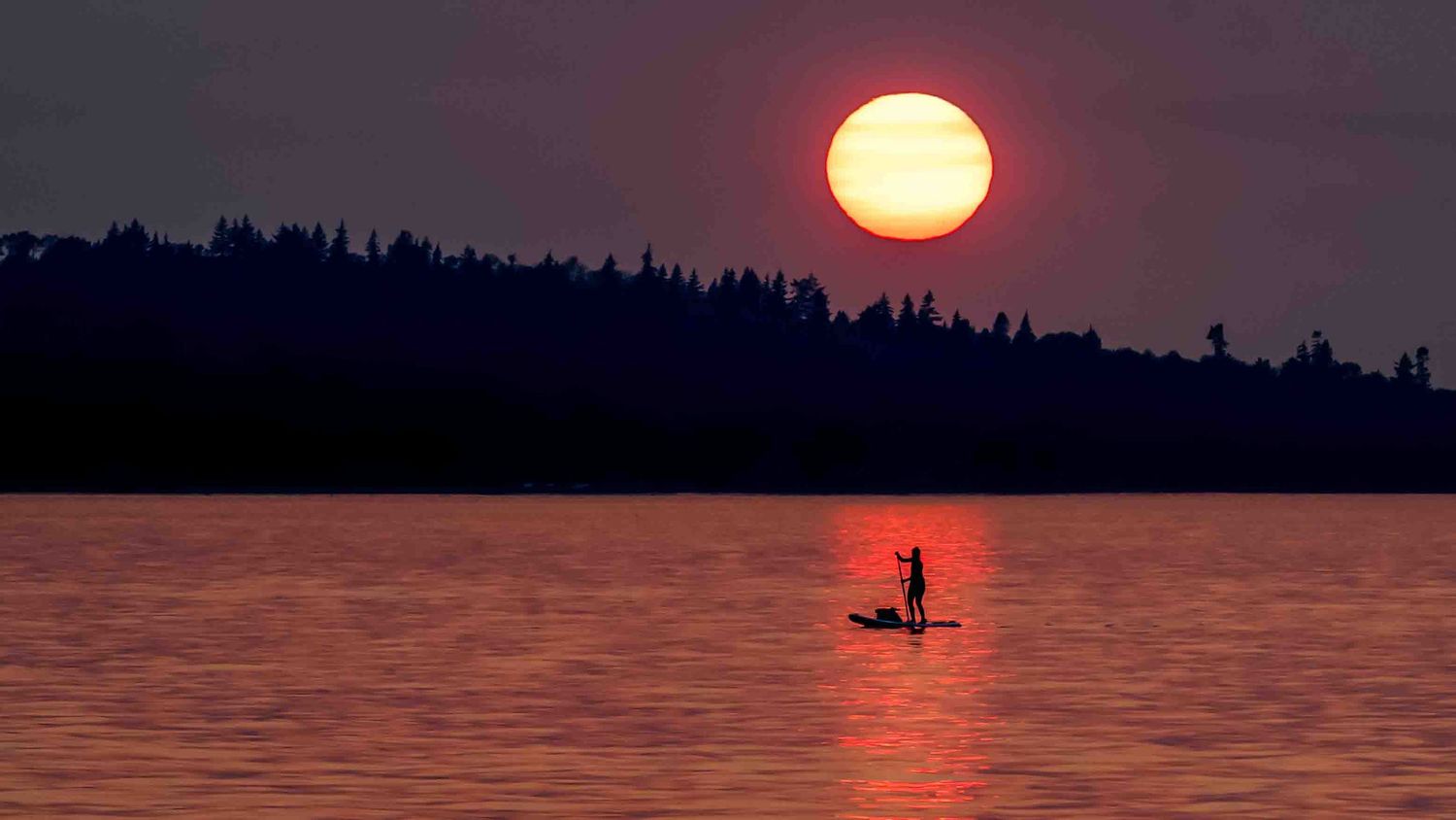 Dramatic sunset over a lake with silhouetted standup paddle boarder, trees and orange sky reflections.