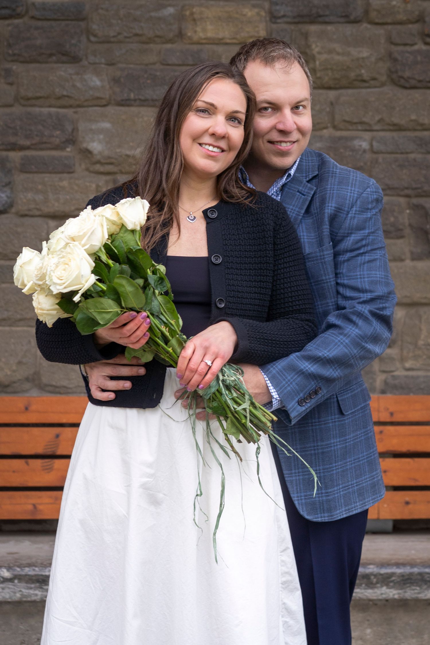 Happy couple posing with white roses bouquet, bride in white skirt and groom in blue suit jacket against stone wall.