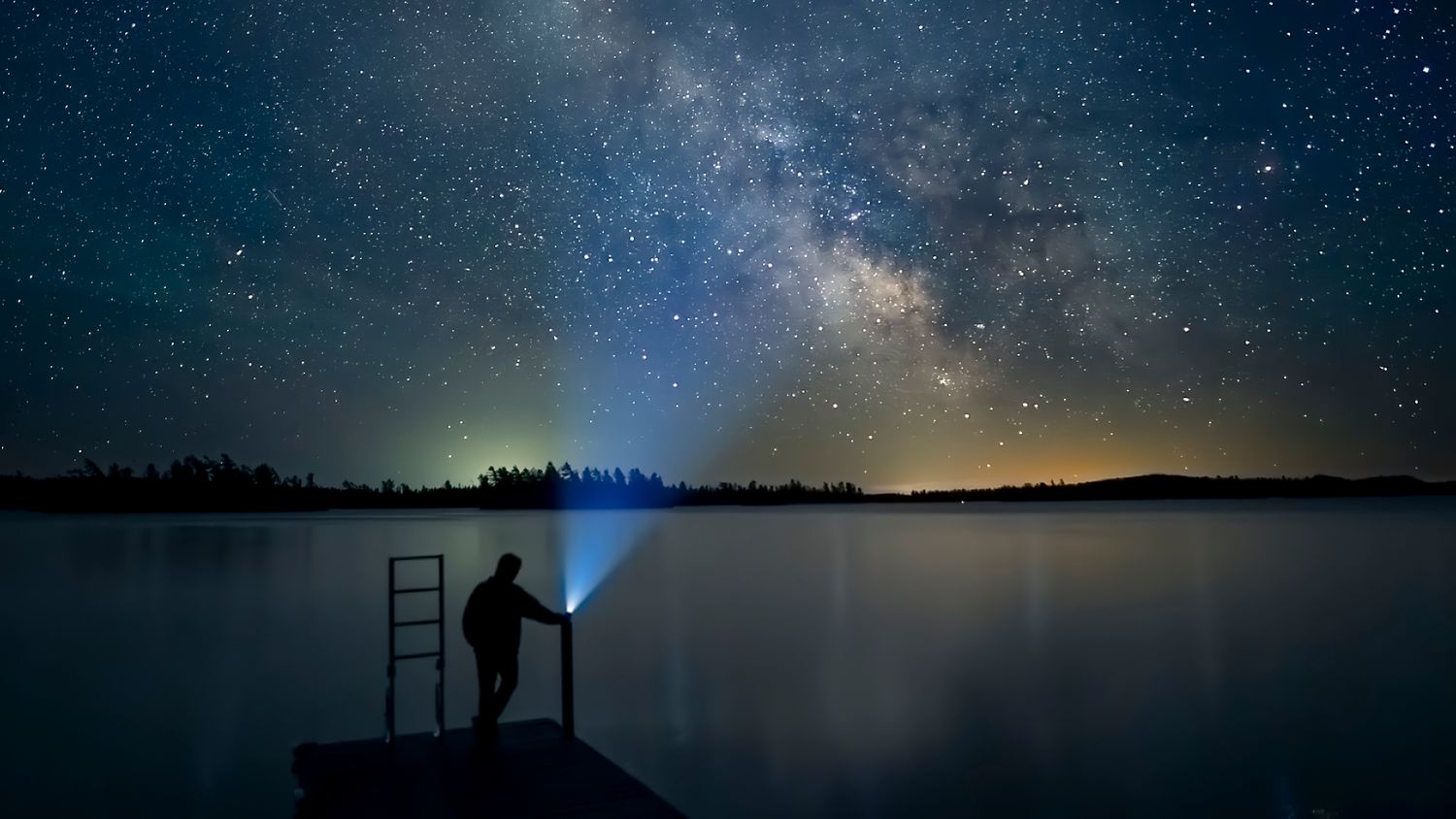 Stunning panorama of the Milky Way galaxy over a dark landscape with silhouetted figure who shines a light back up towards the galaxy