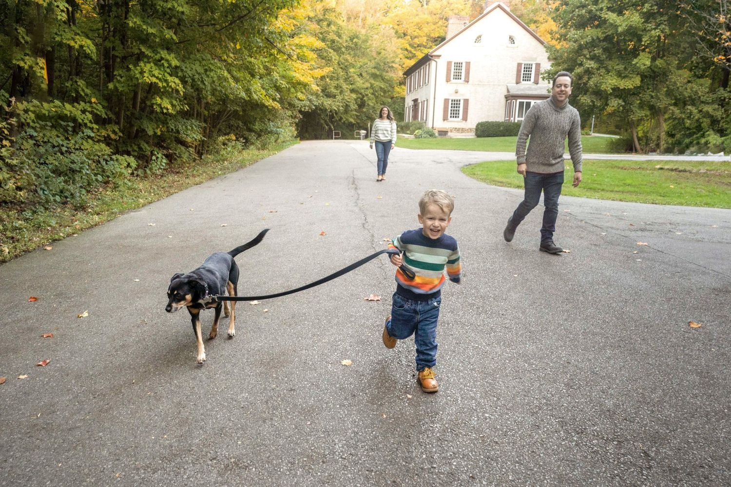 A young boy runs with his dog on a leash, with parents in the background