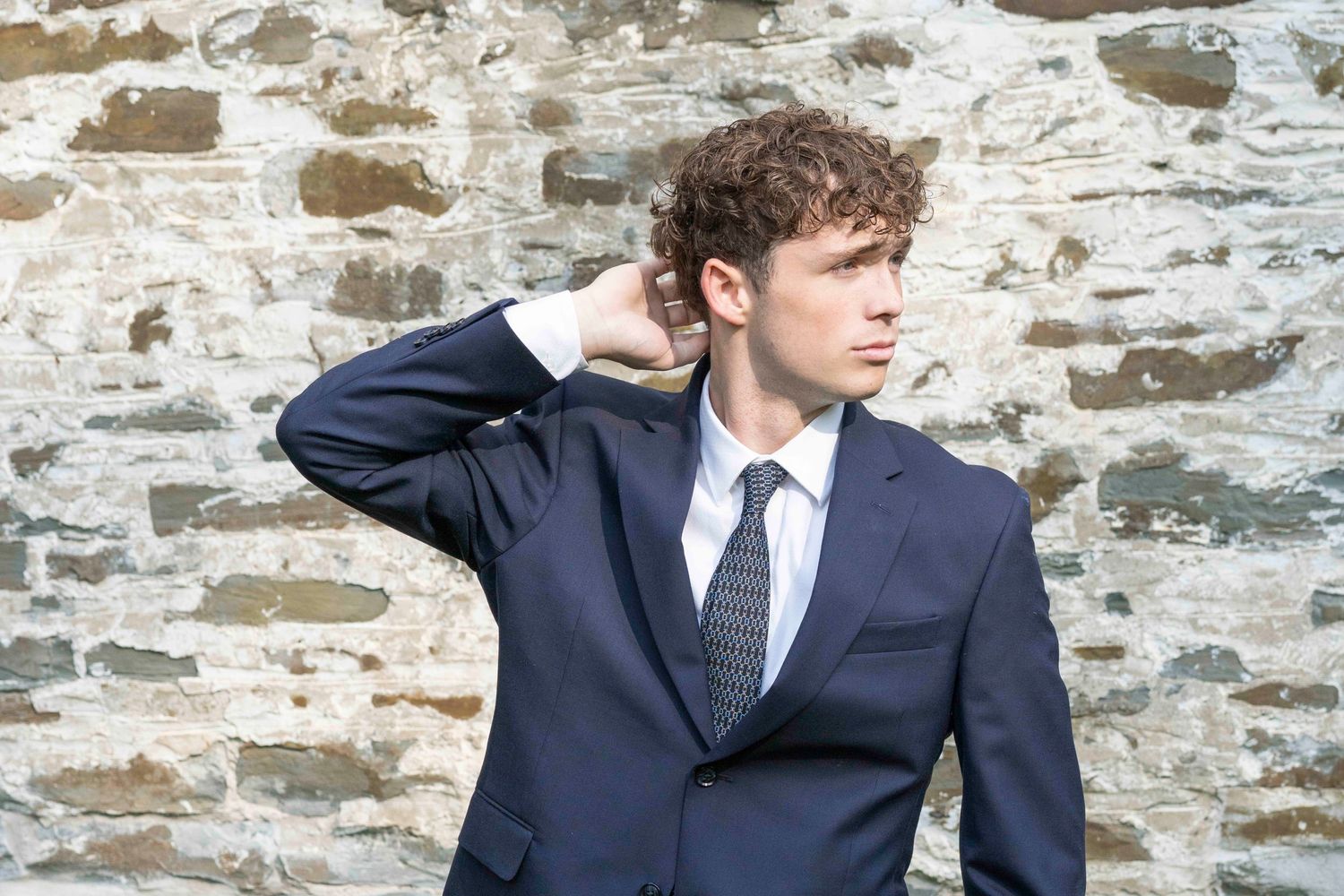 A person in a navy suit and tie stands thoughtfully against a rustic stone wall background.