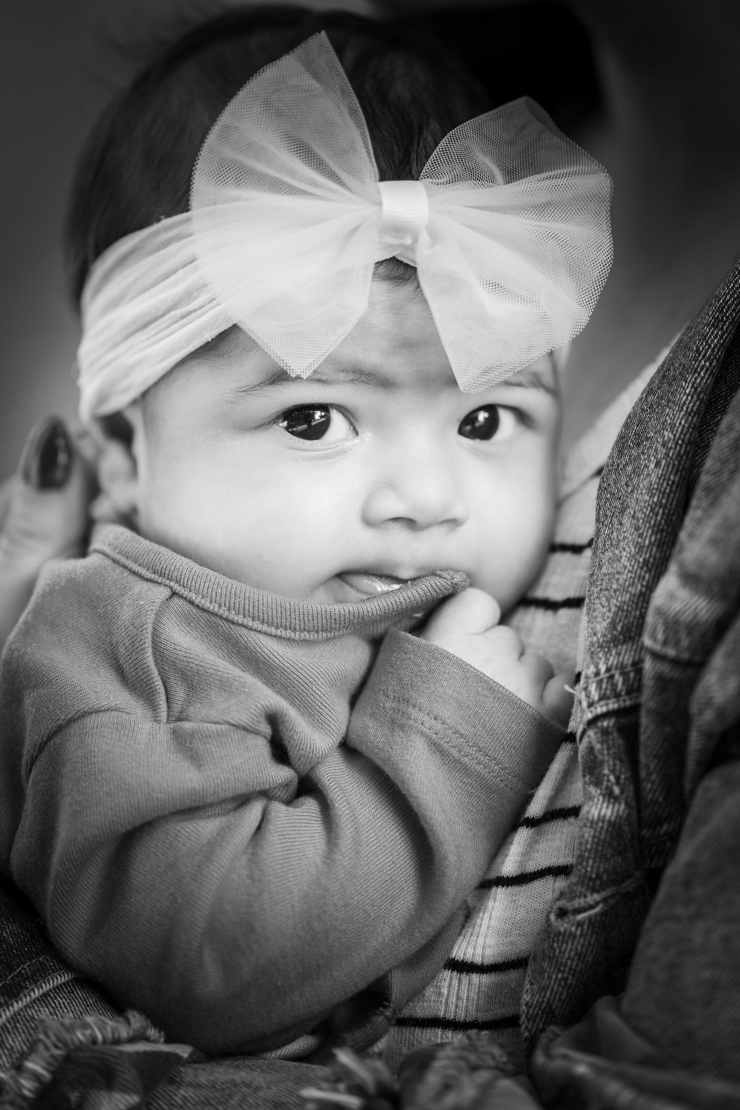 A black and white close-up portrait of a young baby girl wearing a white bow headband and looking directly at the camera.