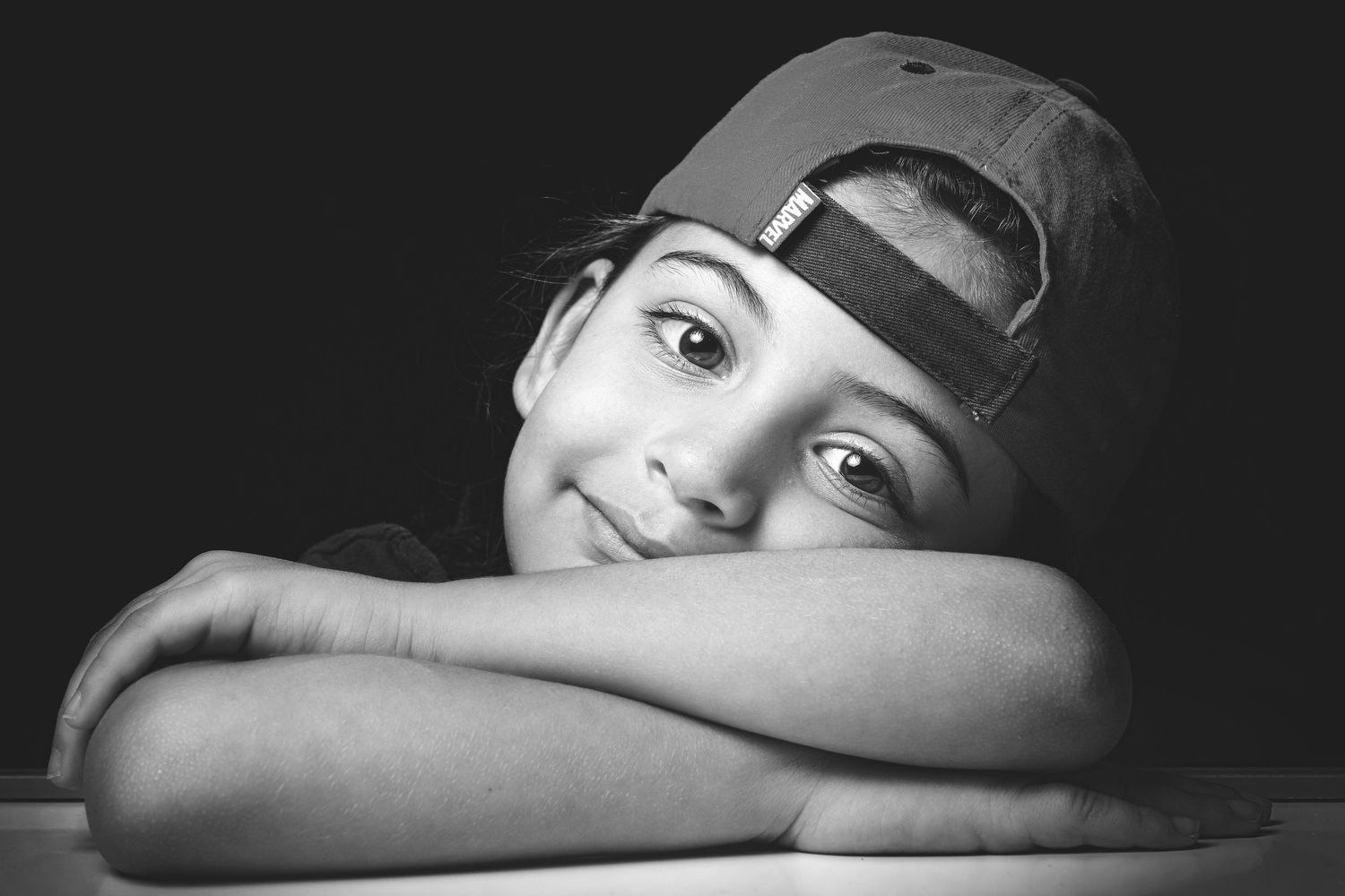 Black and white portrait of a smiling child wearing a backwards cap, resting chin on folded arms.