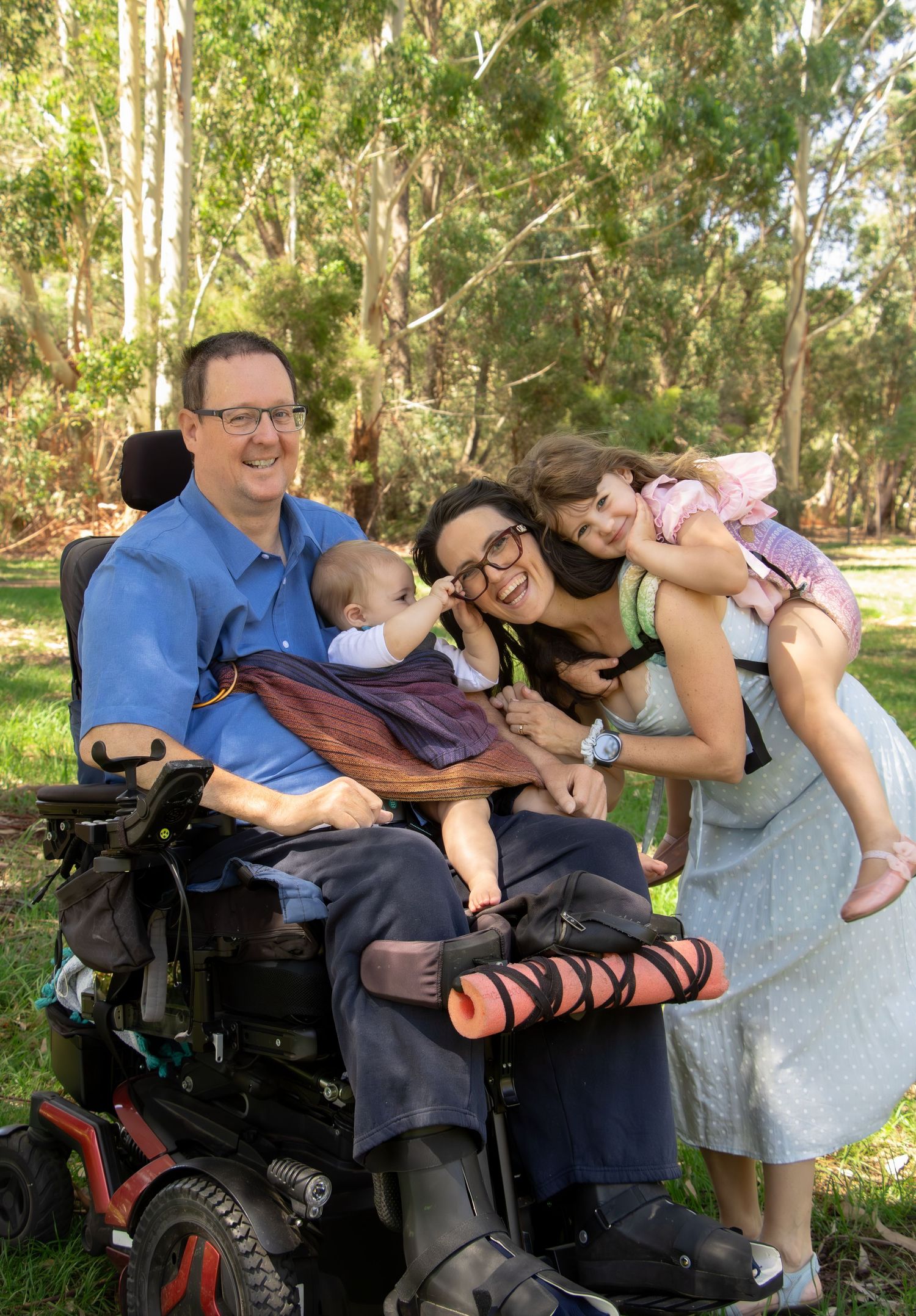 Happy family with man in power wheelchair, woman holding two young children, smiling outdoors in a lush green park setting.