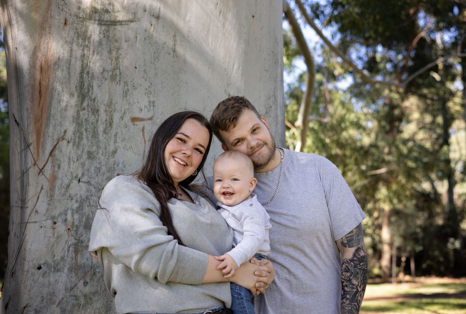 Happy young family with tattooed father, mother, and laughing baby posing outdoors against a large tree.