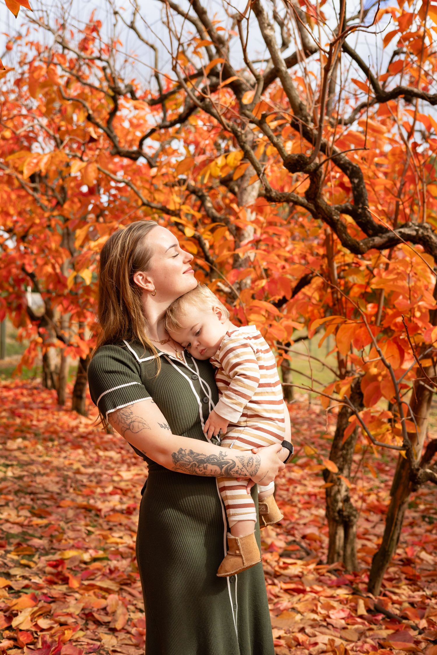 Mother holding sleeping toddler among vibrant red autumn trees, wearing green dress with tattoos visible.