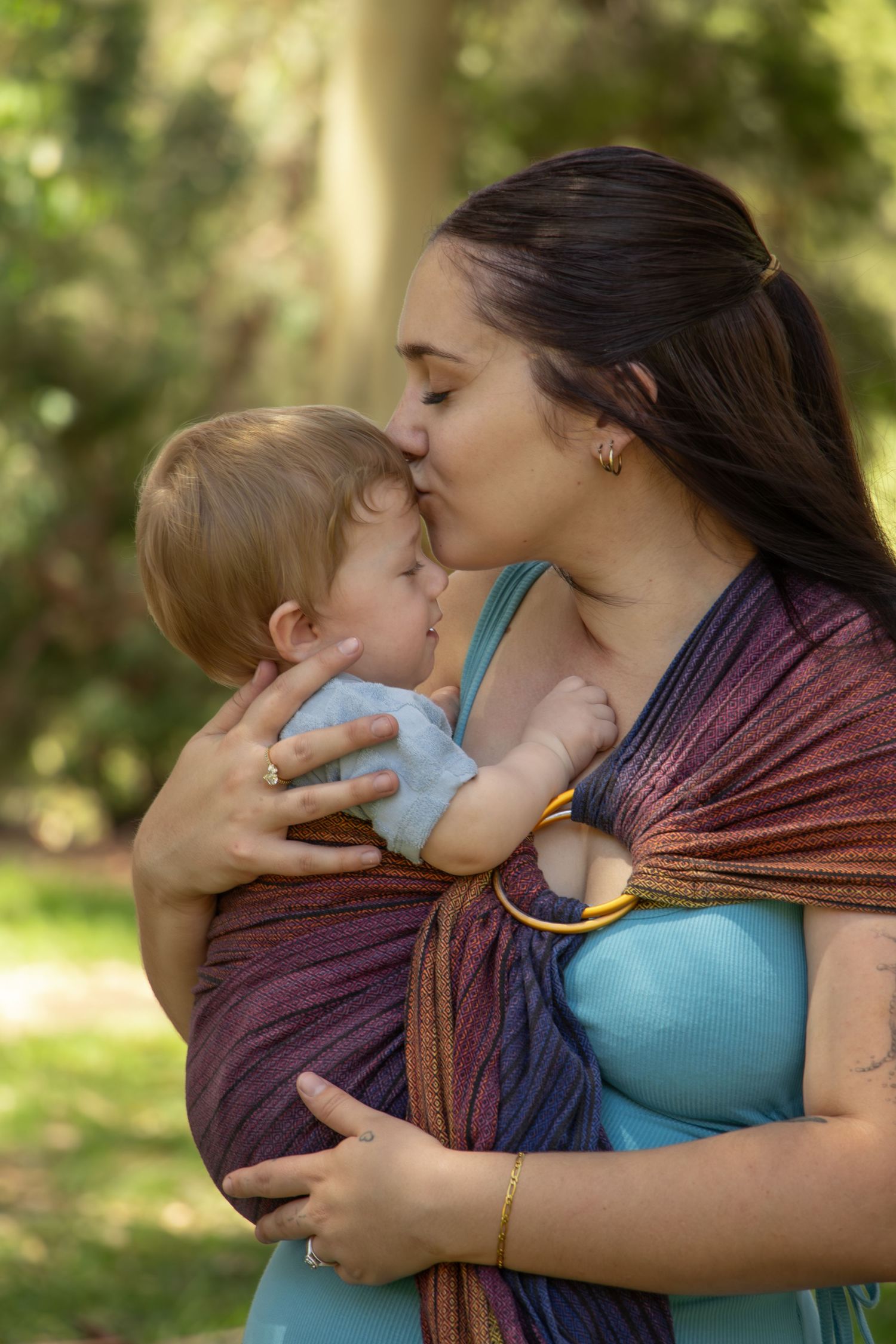 Mother kissing baby in colorful ring sling carrier outdoors in green park setting.