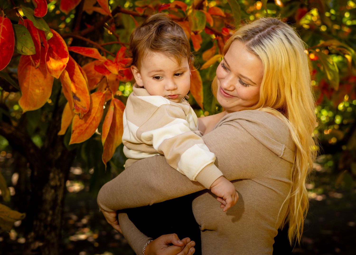 A smiling blonde woman holds a young toddler boy among vibrant red and orange autumn leaves outdoors.