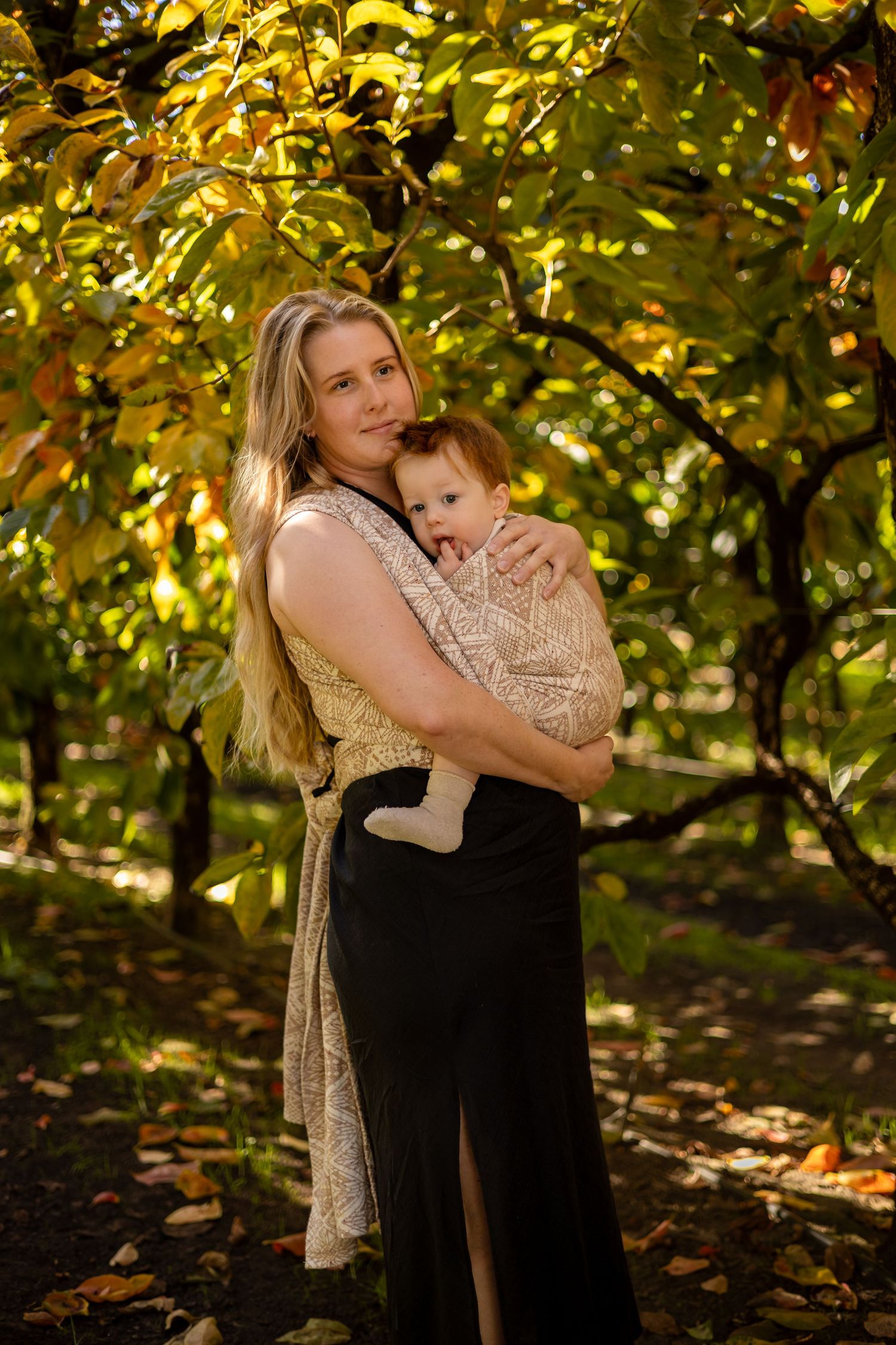 A blonde mother tenderly holds her baby in a sunlit autumn orchard, both dressed in warm tones.