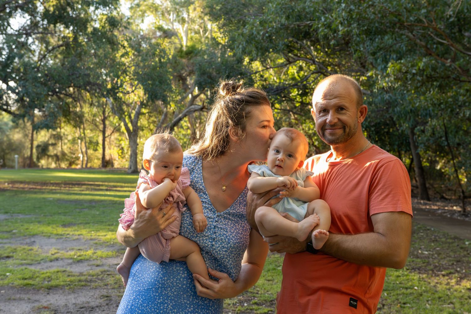 A smiling couple holds two babies in a sunny park with lush green trees in the background.