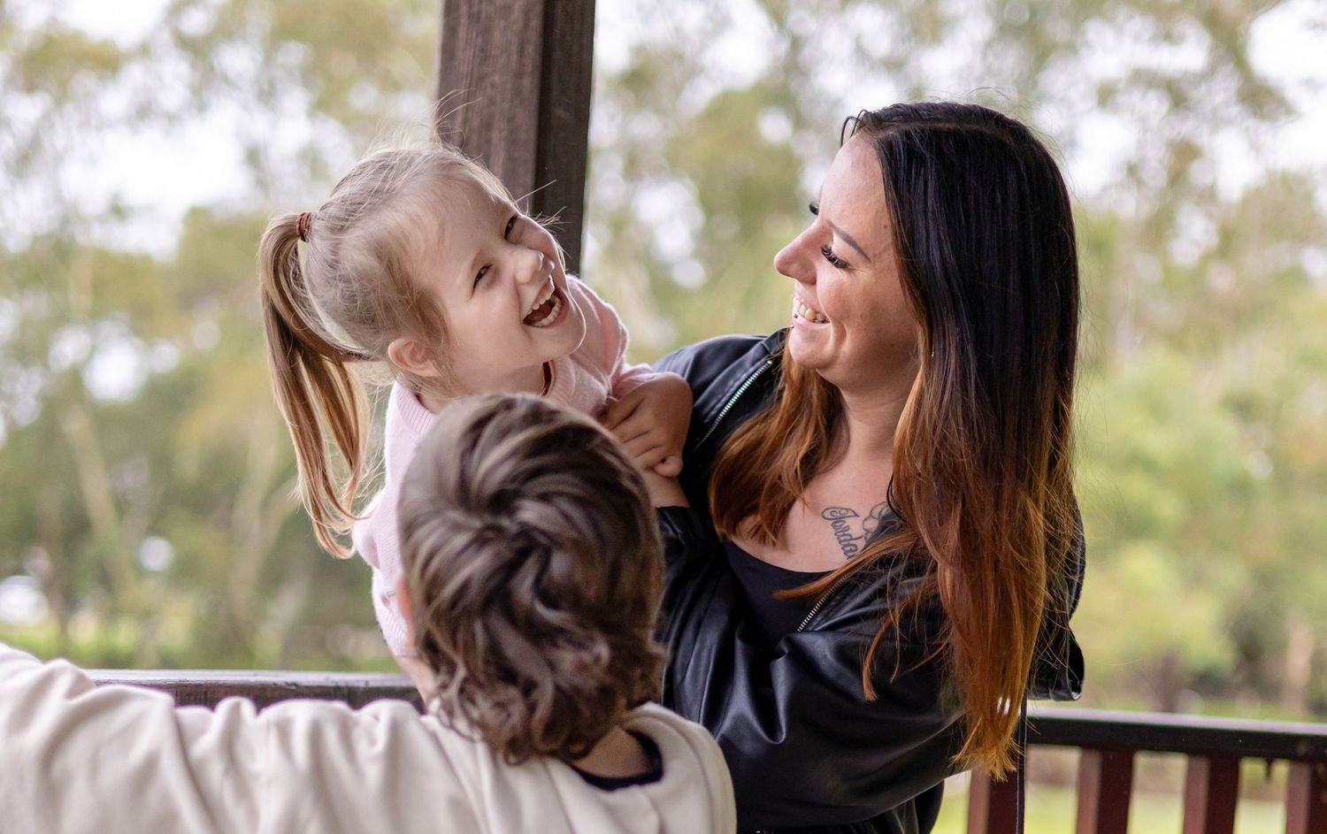 A woman smiles while holding a laughing little girl on an outdoor deck surrounded by lush greenery.