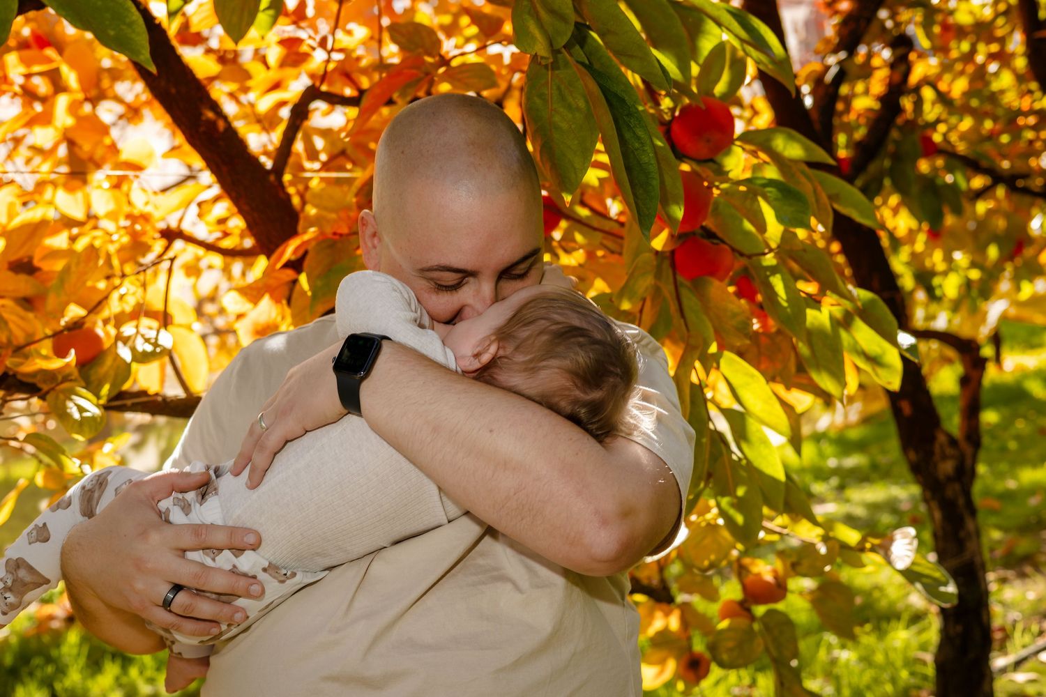 Bald man tenderly hugging a child outdoors among vibrant autumn orange and yellow foliage trees.