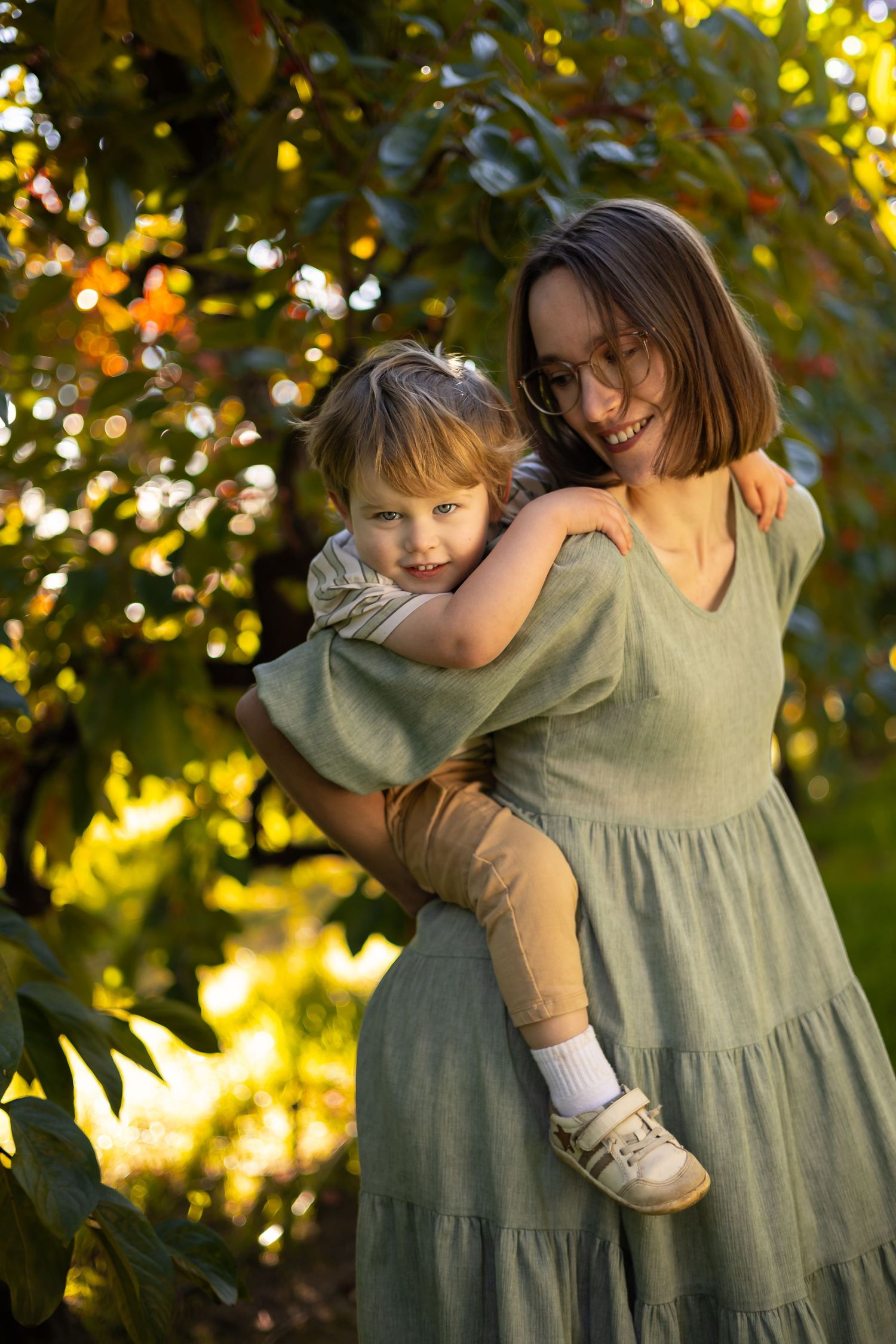 A smiling woman in a green dress holds a young child outdoors among sunlit autumn trees.