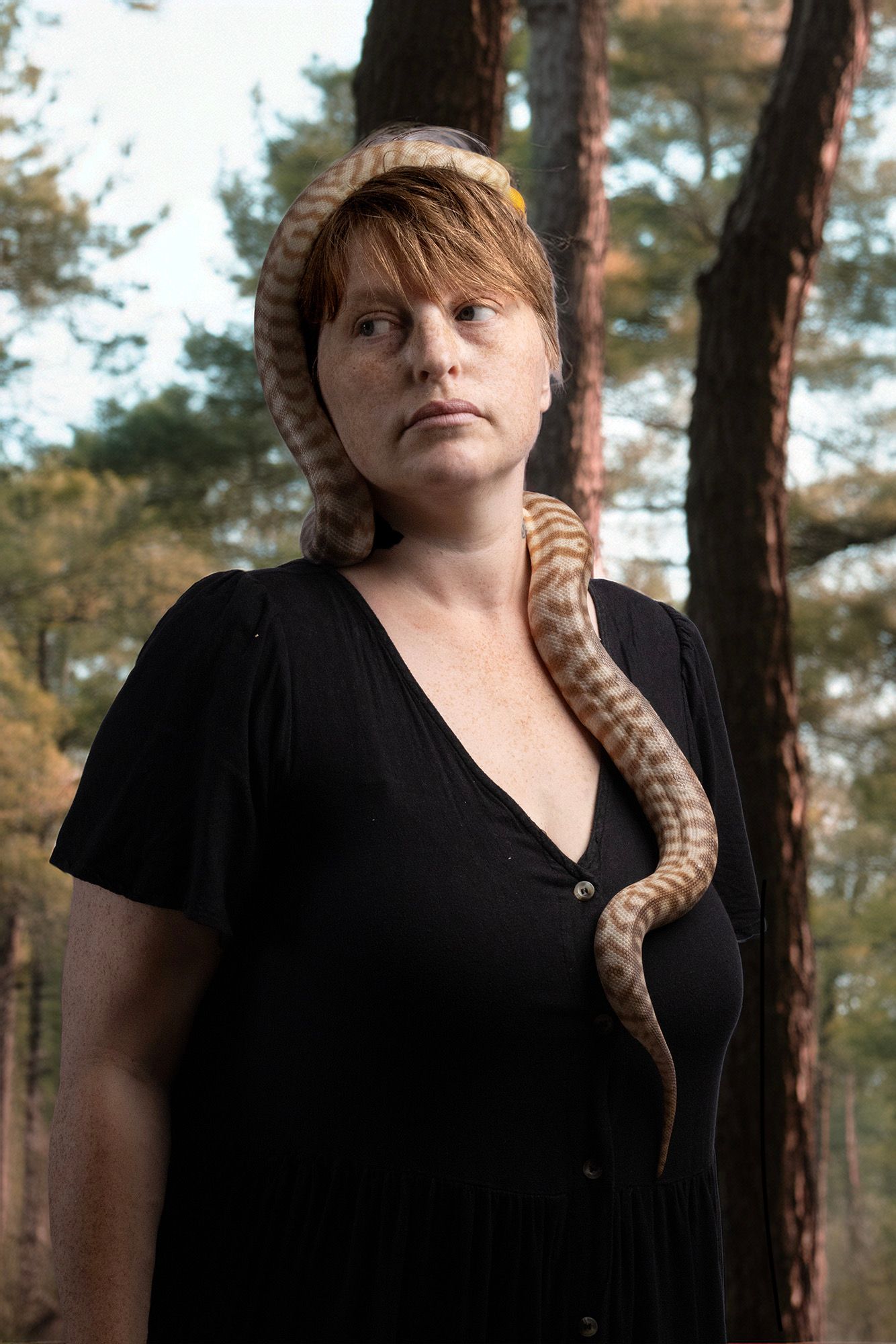 Woman in black dress with a snake draped over her head and chest, standing in a forest.