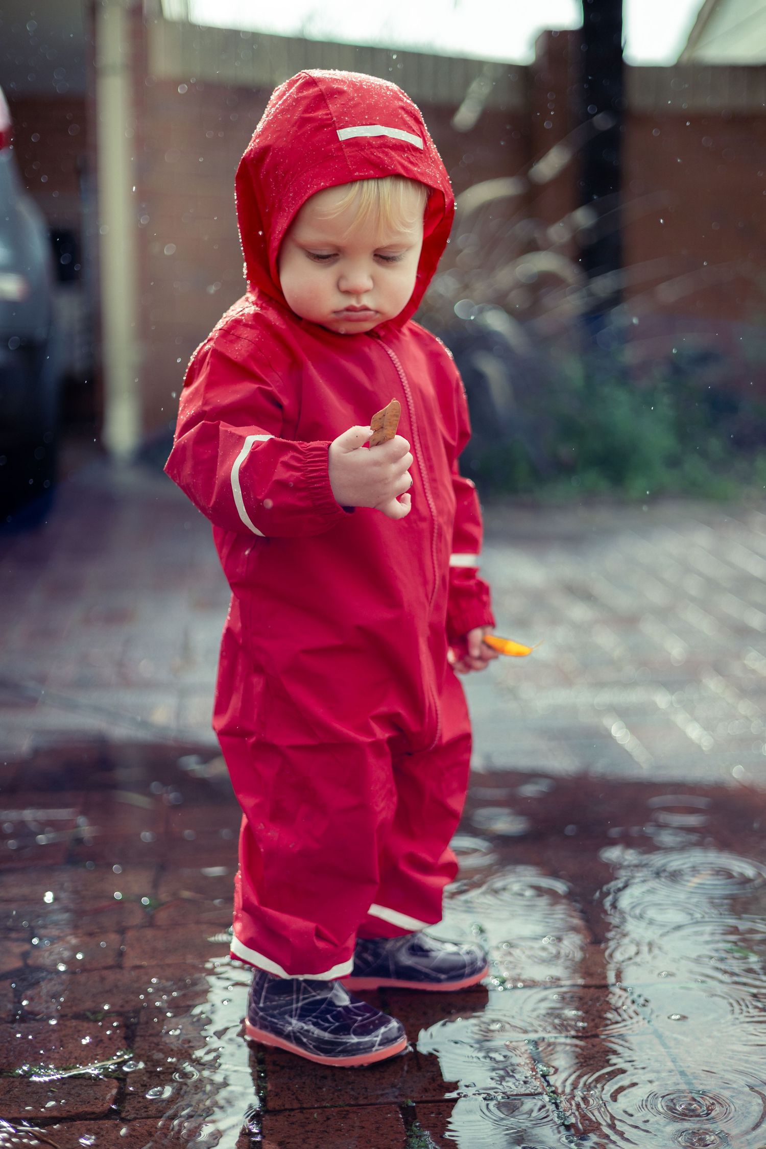 Toddler in red rainsuit and boots splashing in puddles on a rainy day outdoors.