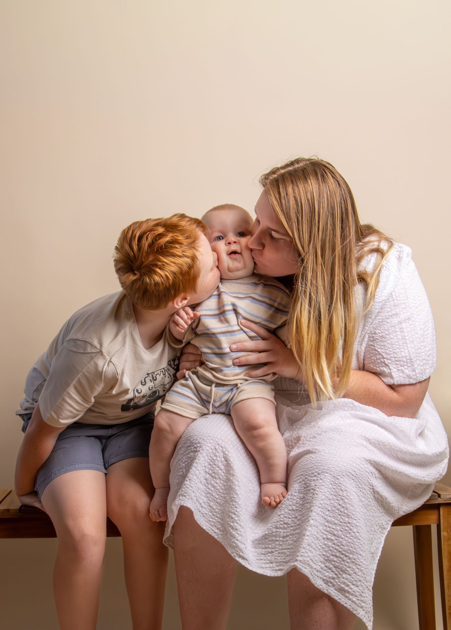 A mother and young redhead child kissing a smiling baby on the cheeks while seated on a wooden bench.