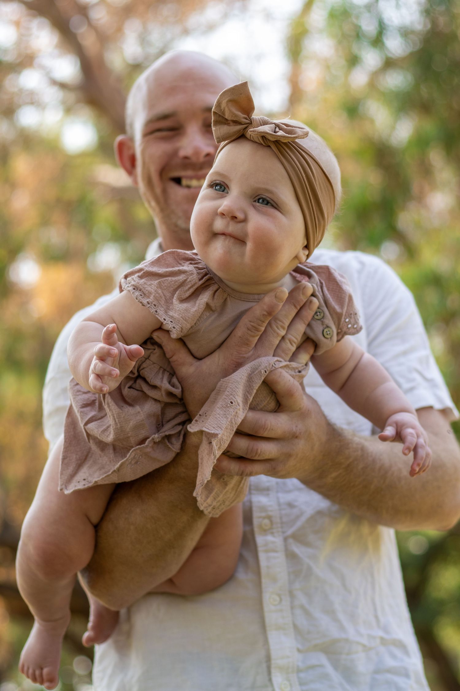 Smiling bald father holding his adorable baby girl in a pink dress and bow headband outdoors.