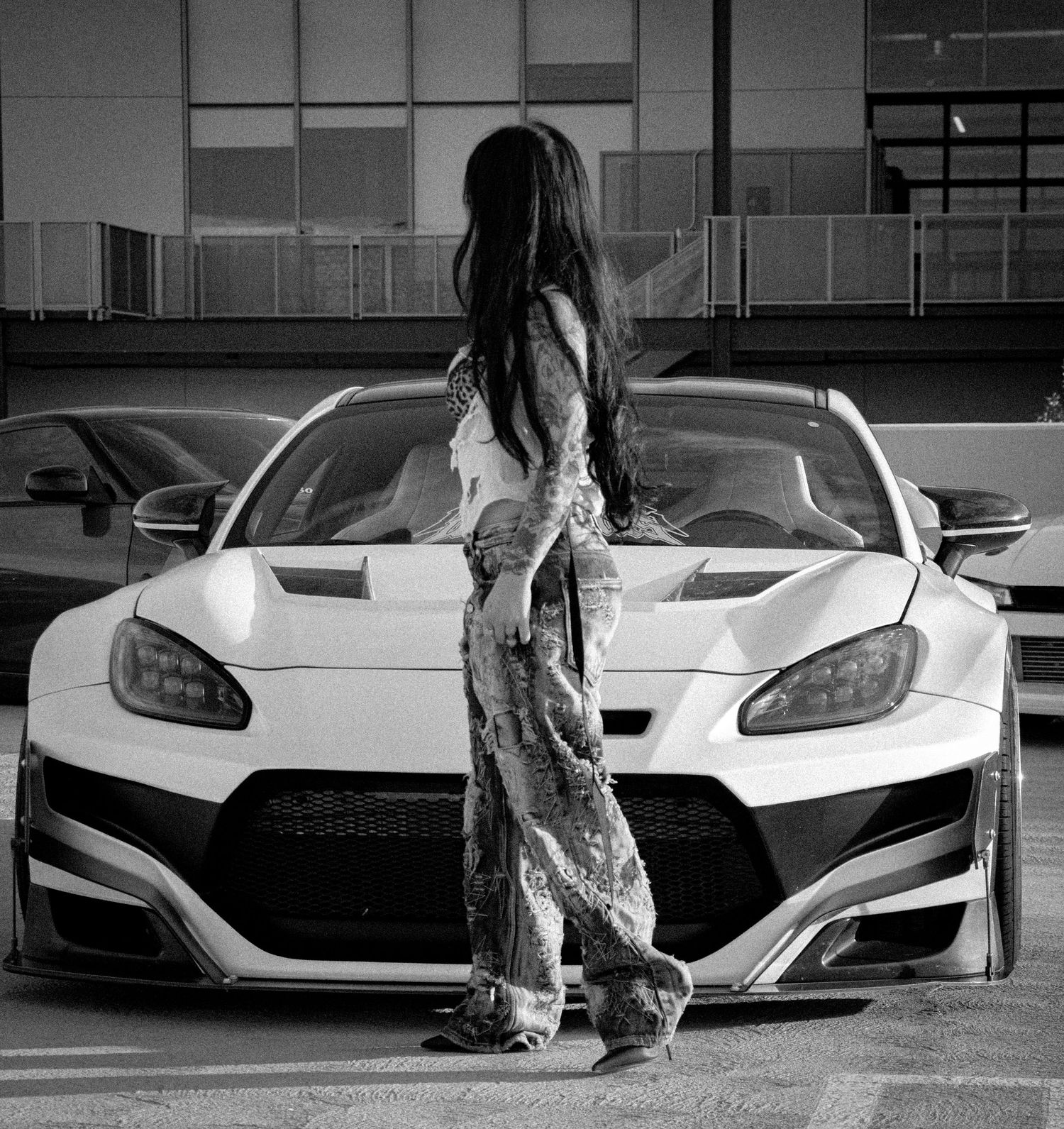 Black and white photo of a woman with long hair standing in front of a wide-body sports car in a parking lot.