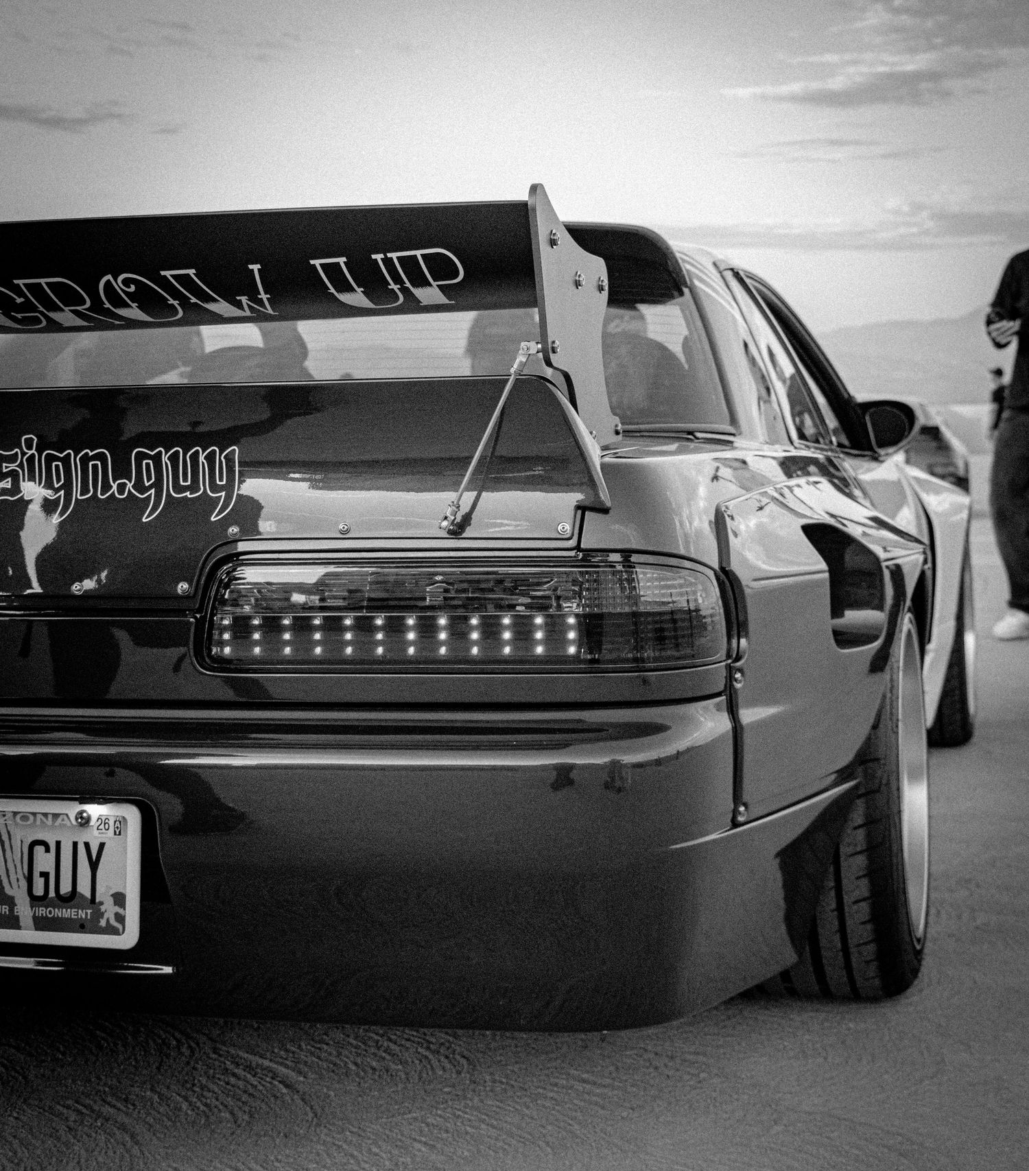 Black and white photo of a modified sports car with GROW UP decal and custom LED taillights at a car meet.
