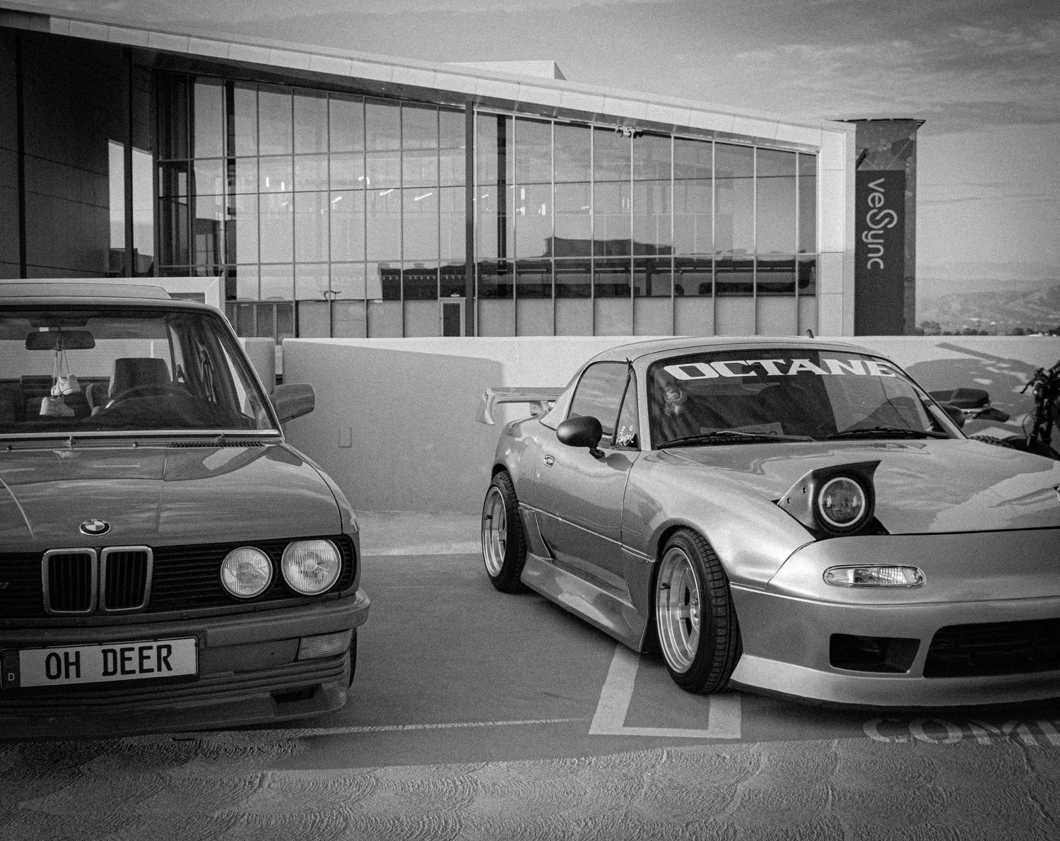 Black and white photo of two modified cars, a BMW E28 and a widebody Mazda MX-5, parked outside a modern building.