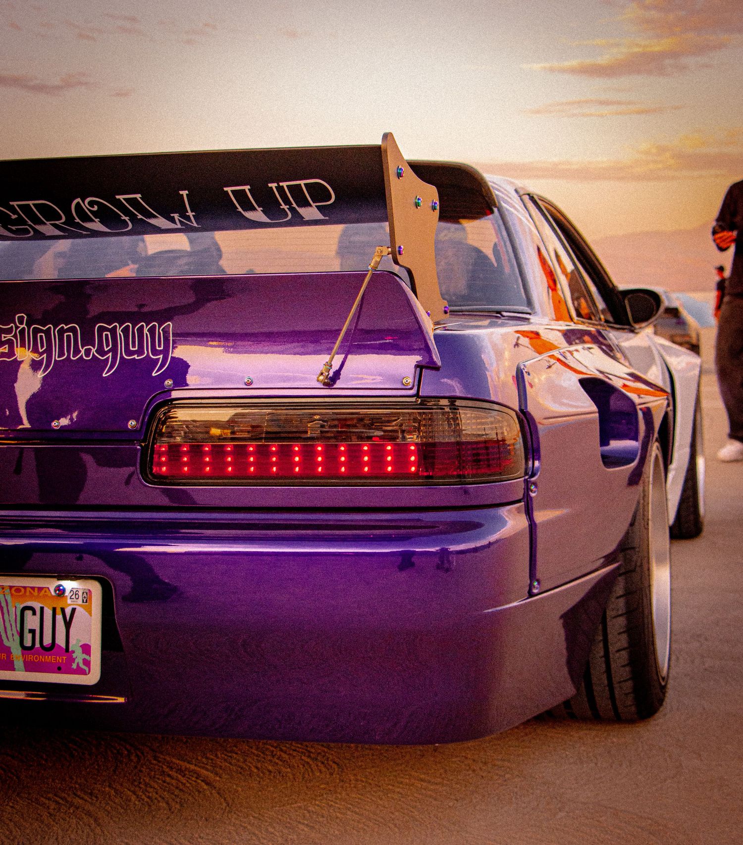 Purple modified sports car with LED taillights, large spoiler, and custom license plate at sunset.