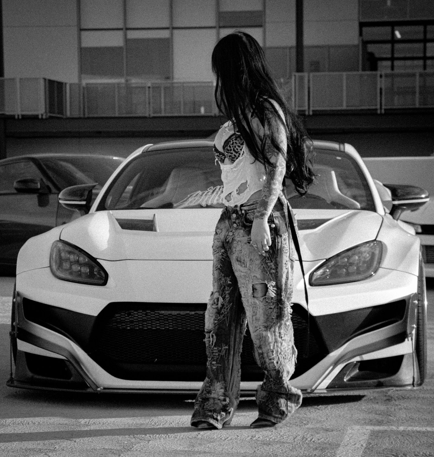 Woman with tattoos poses in front of a white modified sports car with wide body kit in a parking lot.