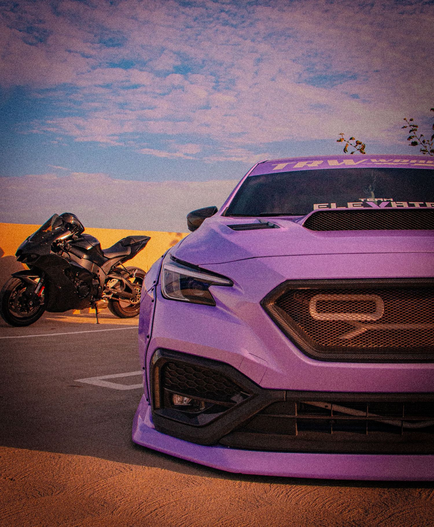 Purple modified Subaru WRX with custom body kit parked next to a sport motorcycle under a cloudy sky.