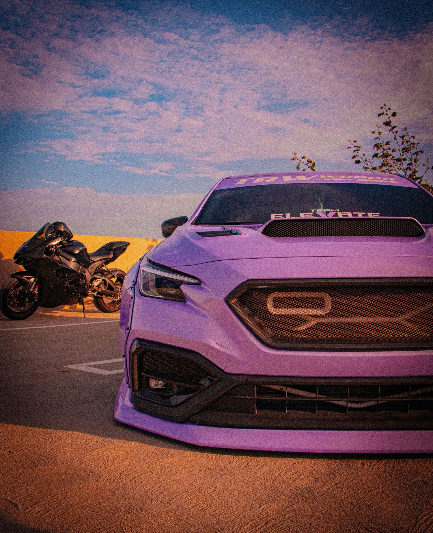 Purple Subaru WRX with custom body kit parked beside a black sport motorcycle under a dramatic sunset sky.