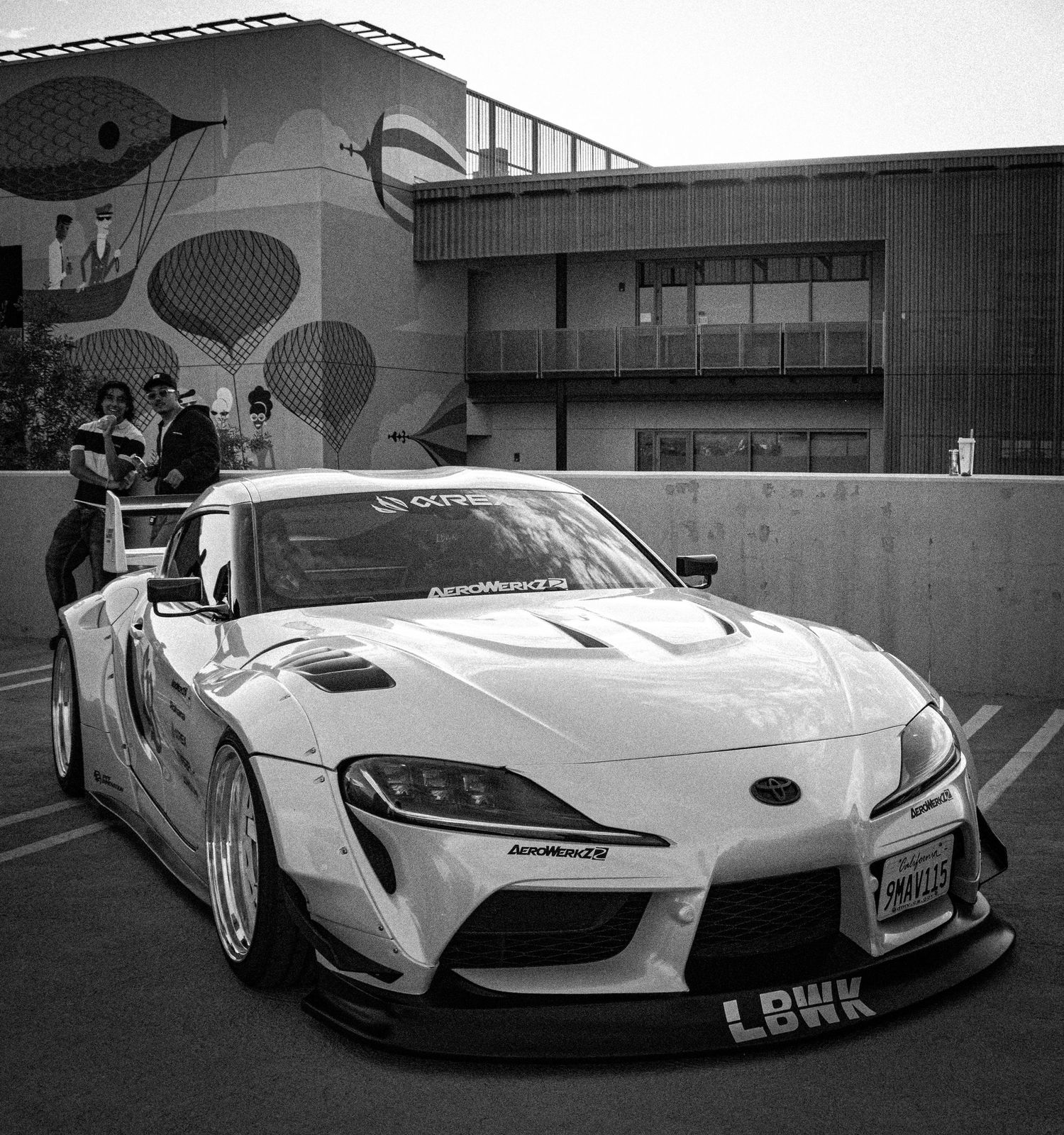 Black and white photo of a widebody Toyota Supra with LBWK kit parked in a lot near a modern building.
