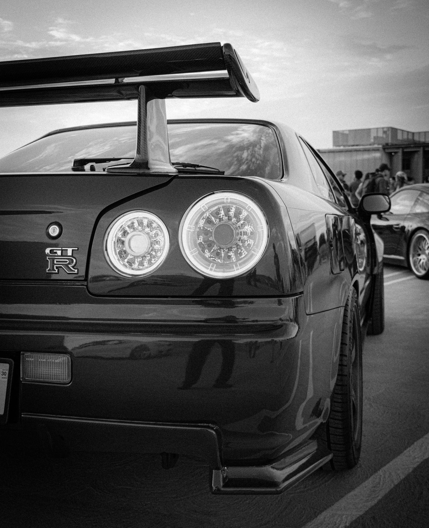 Black and white rear view of a Nissan GT-R R34 with iconic round taillights and large rear spoiler at a car show.