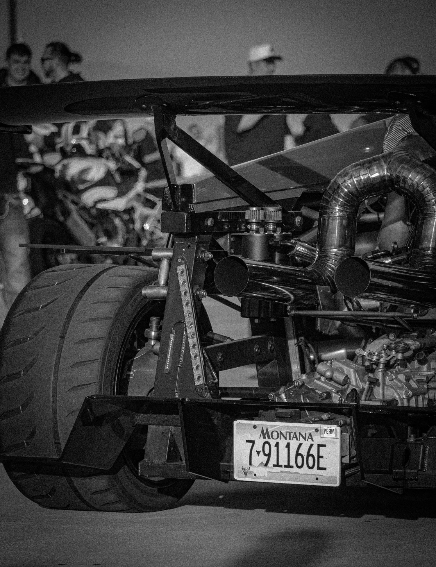 Black and white photo of a Montana-plated race car rear view showing large tire, roll cage, and exposed engine components.