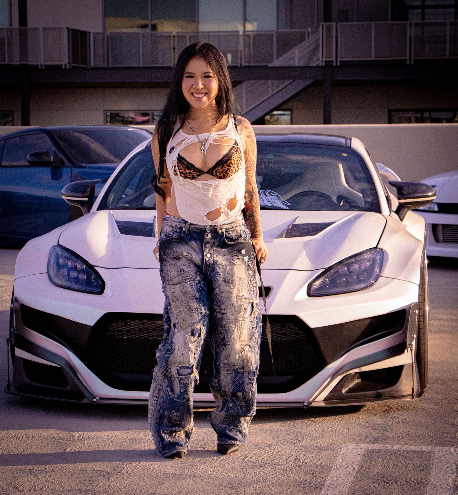 Young woman in ripped jeans and crop top poses in front of a white custom widebody sports car in a parking lot.