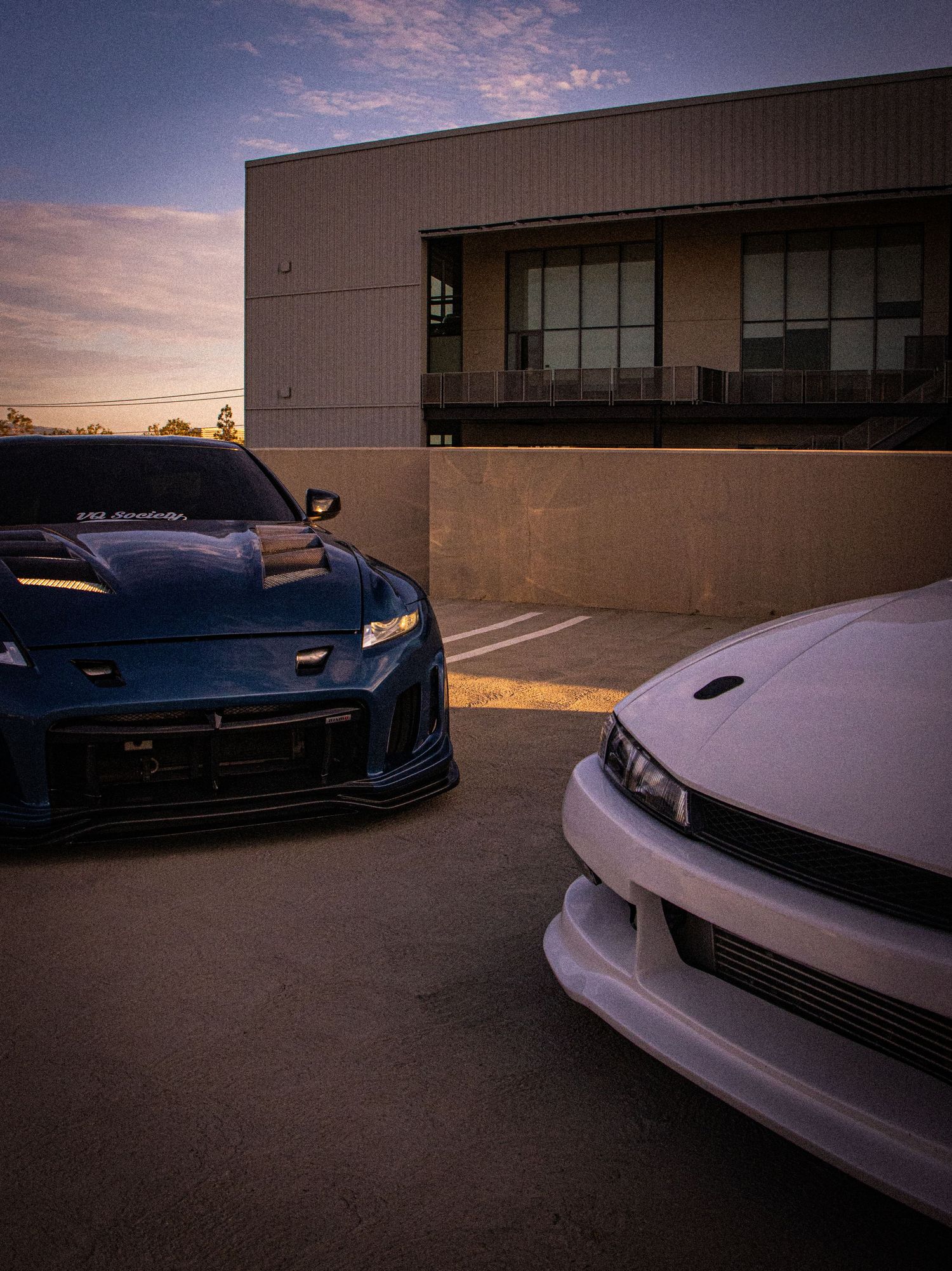 Two modified sports cars, a blue and white, parked at dusk near a building with dramatic sky.